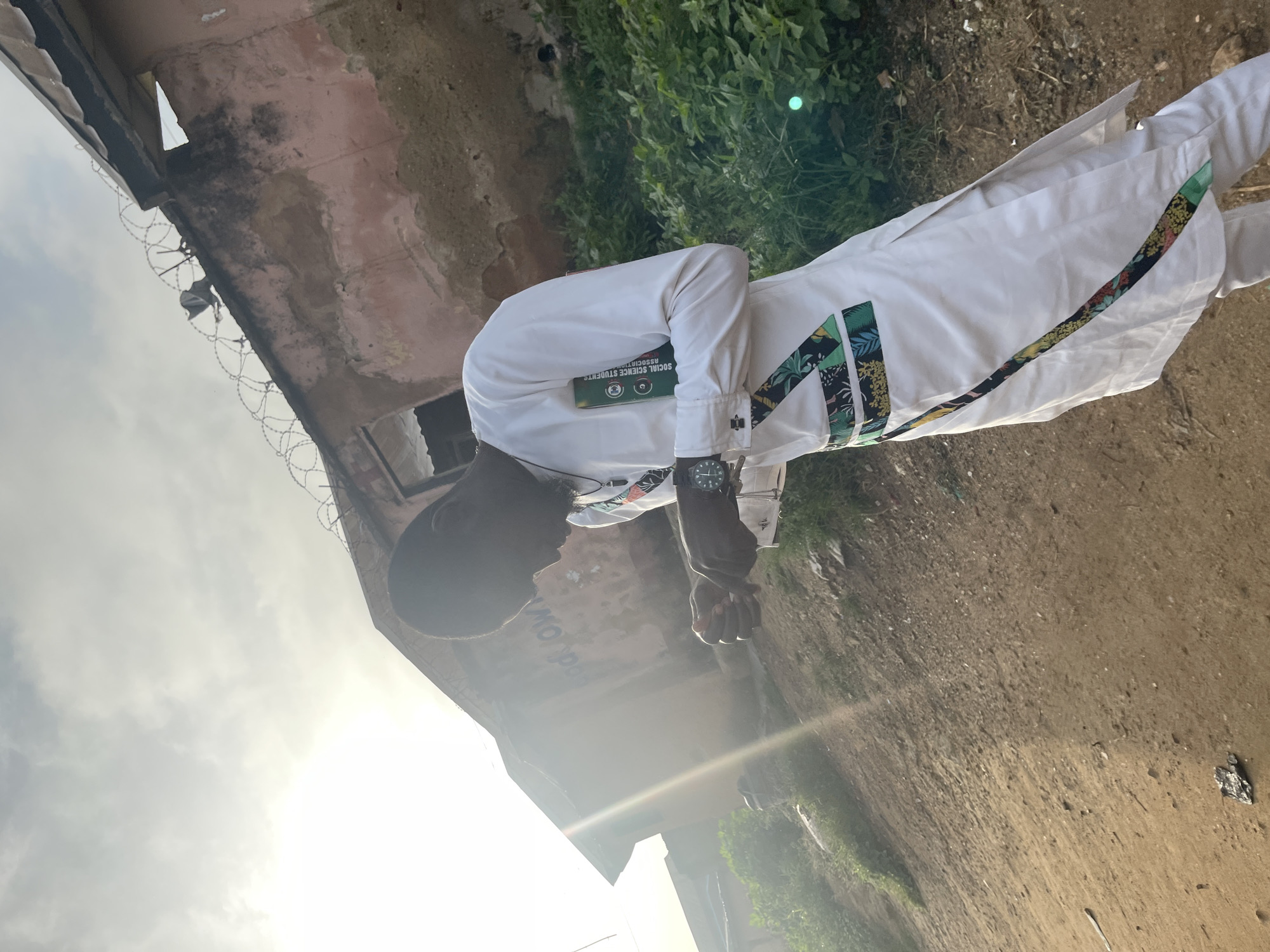 A young Black male, dressed in a white traditional outfit adorned with colorful geometric patterns, stands outdoors in what appears to be Rafin Par, Nigeria. He is looking down at his hands, which are clasped in front of him. A watch is visible on his left wrist. The background features a dilapidated building with a pinkish-red exterior, topped with barbed wire. Overgrown green vegetation surrounds the building and the dirt ground. The sky is overcast with a bright haze, suggesting either early morning or late afternoon light. The overall atmosphere is one of poverty and neglect.