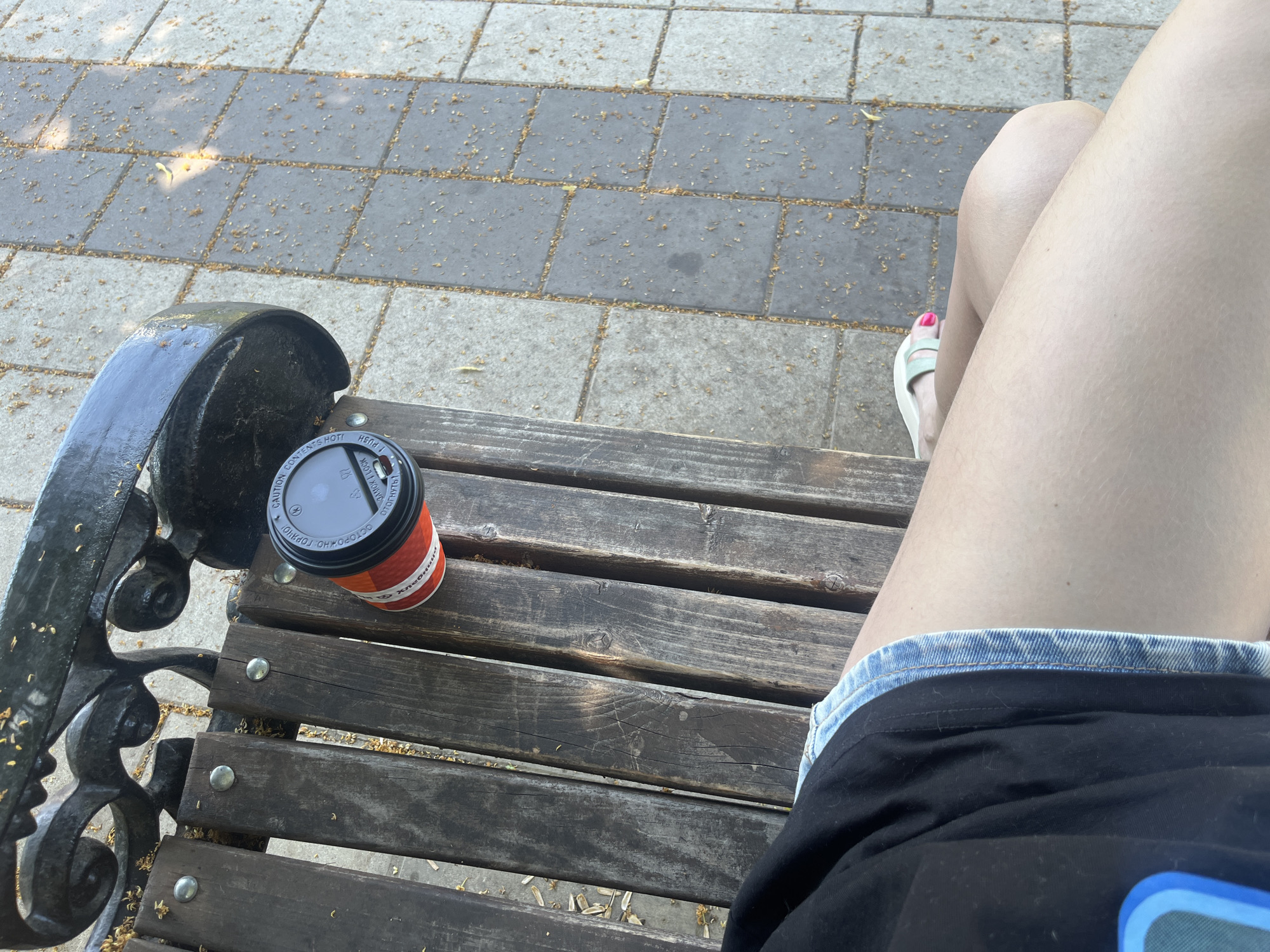 A person is sitting on a wooden bench in an outdoor setting. A red and white disposable coffee cup with a black lid rests on the bench. The person is wearing a black t-shirt and denim shorts. Their right foot is visible, wearing a light-colored sandal with a pink toenail. The ground next to the bench is paved with gray and beige square tiles, and there are scattered yellow petals on the ground. The scene appears to be a park or a public seating area.