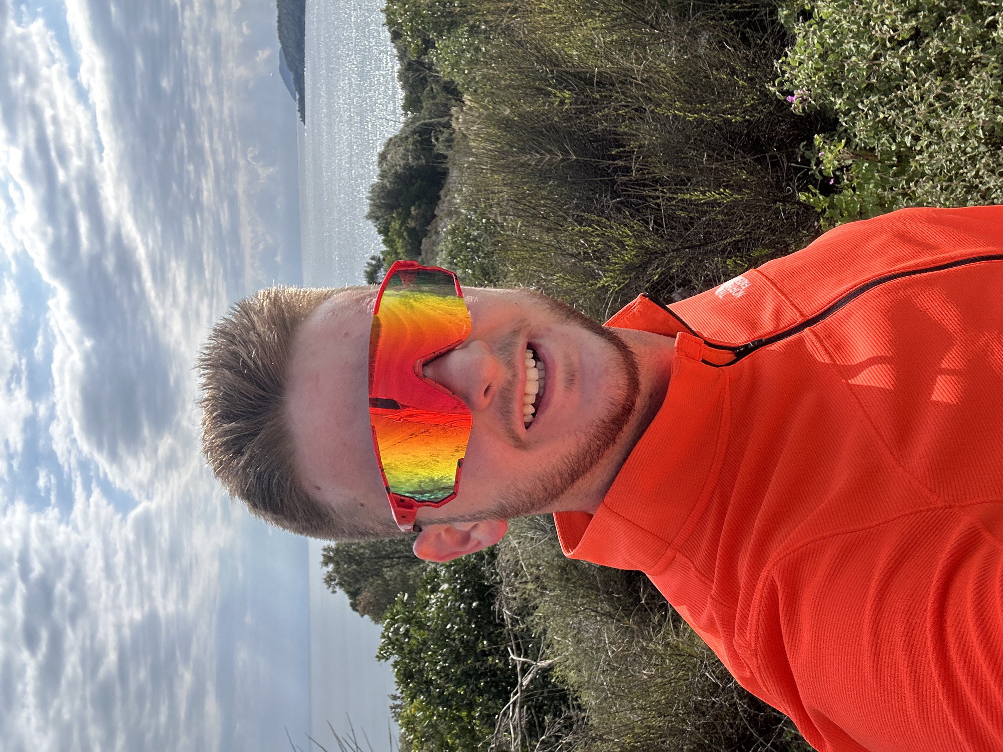 A man is taking a selfie outdoors in Ολυμπιάδα, Greece. He is wearing an orange shirt and brightly colored sunglasses, and smiling. The background shows a body of water, possibly the sea, reflecting the cloudy sky. There are bushes and trees on the right side of the image.