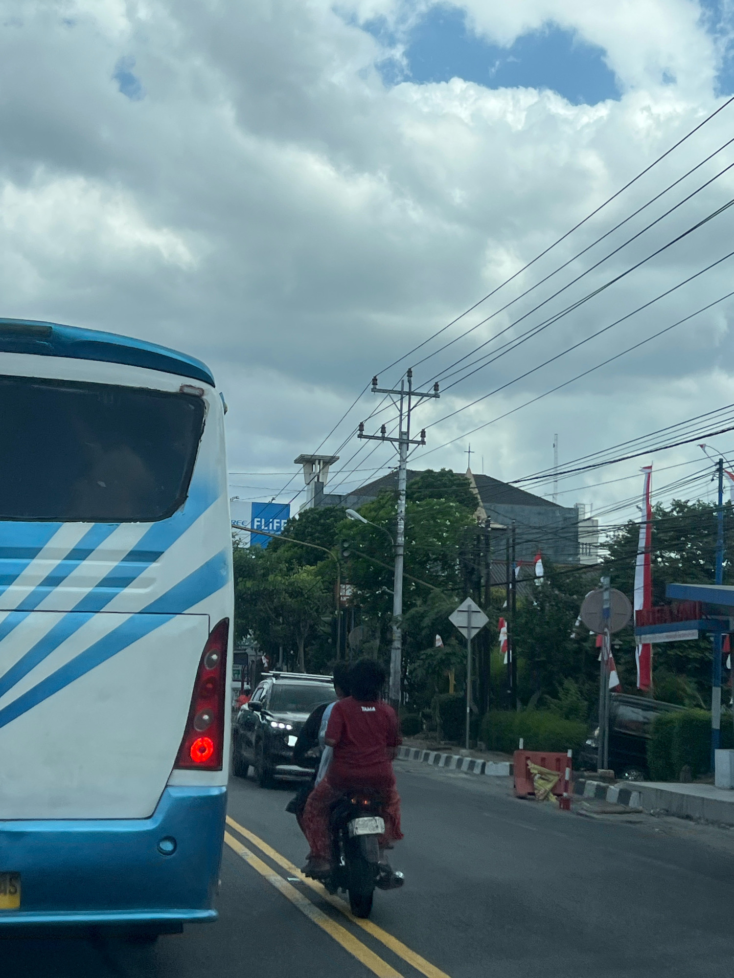 In Magelang, Indonesia, on a cloudy day, traffic moves along a road. A large white and blue bus is prominently featured on the left, its rear end filling much of the frame. A motorcycle carrying two individuals is in the lane next to the bus, with the rider wearing a red shirt and the passenger in a red shirt and patterned pants. A dark SUV is in the distance, also on the road. The surroundings include trees, buildings with visible roofs, and utility poles with numerous wires. Decorative flags, red and white, are visible along the roadside. A construction barrier with orange and yellow elements is present on the sidewalk. The road is marked with yellow double lines.
