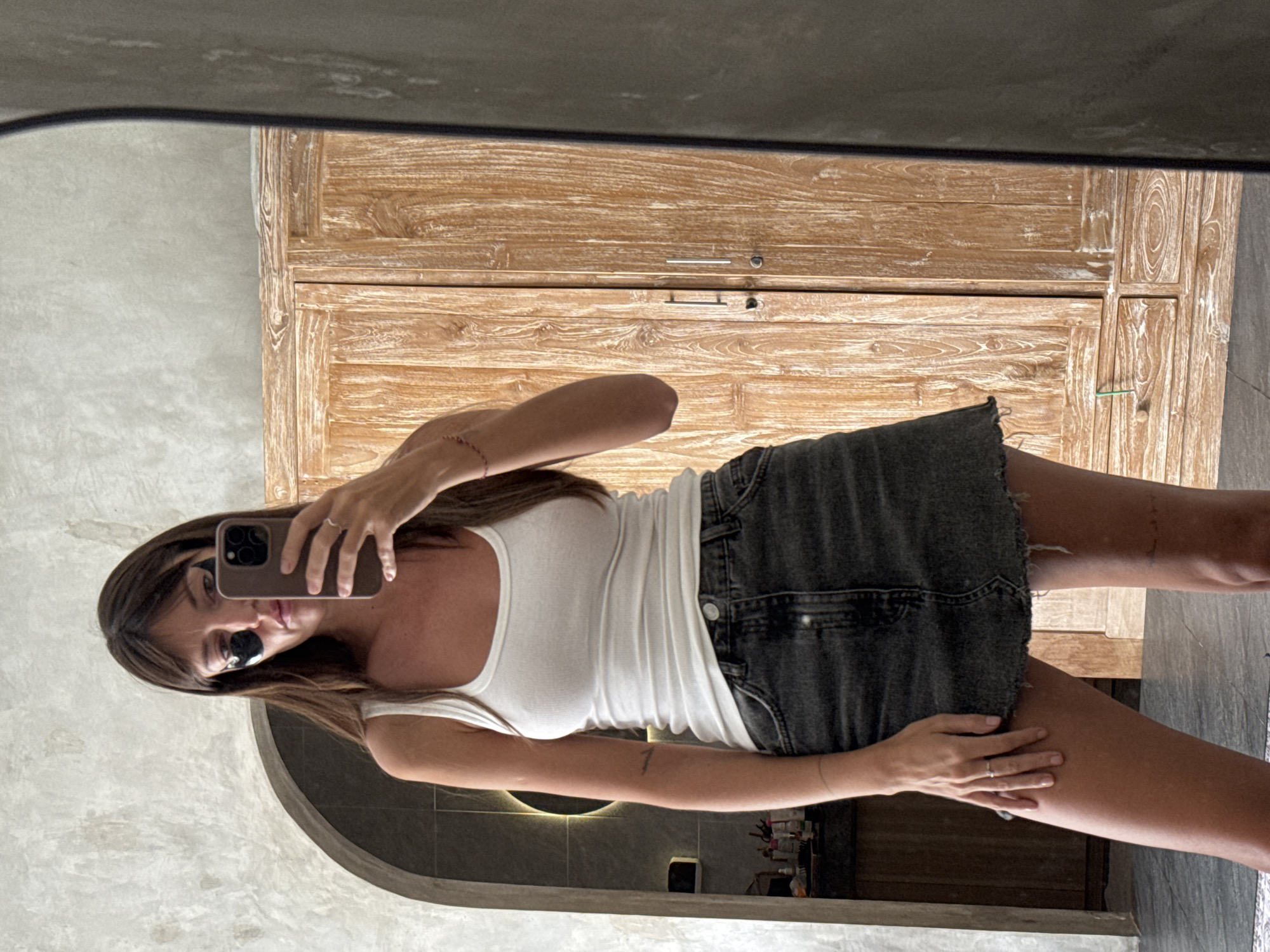 A woman with long brown hair, wearing black eye patches, a white tank top, and a dark denim skirt, stands in what appears to be a bathroom or dressing area and takes a mirror selfie with her phone. She is wearing sunglasses and several rings. The background shows a textured wall, an arched doorway with a circular mirror, and a light-colored wooden cabinet with two doors and drawers. The floor is tiled. The overall aesthetic suggests a modern, possibly spa-like environment.