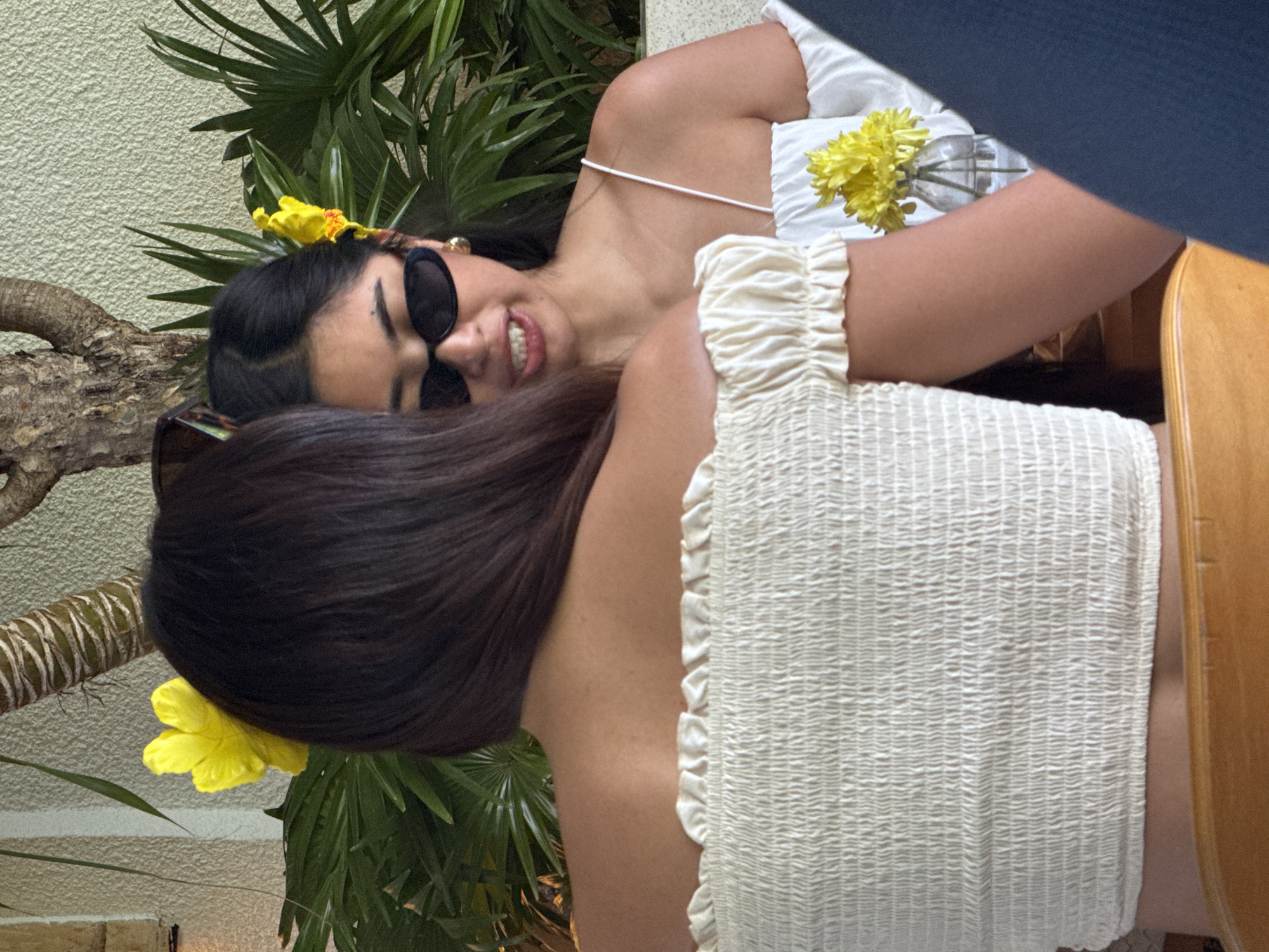 The image depicts two women with dark hair, one wearing sunglasses and a white dress with yellow flowers in her hair and the other in a white smocked top, posing outdoors. They are positioned against a backdrop of tropical plants and a textured wall. A wooden tree trunk-like object is also visible. A glass vase holding yellow flowers is on the table. This scene likely takes place in North Kuta, Indonesia, based on the request context.