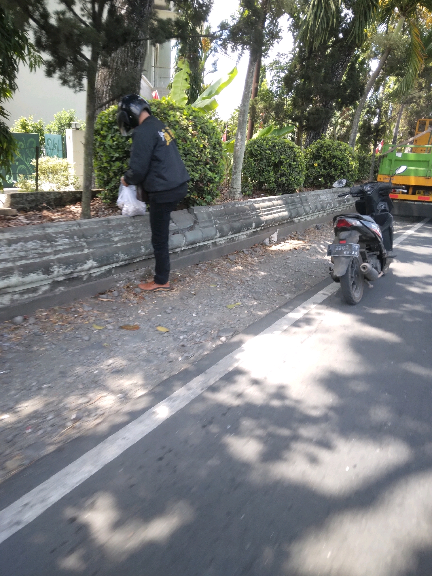 In the city of Sokaraja, Indonesia, a person wearing a black jacket, black pants, and a black helmet is standing next to a motorcycle. The person is holding a white plastic bag in their right hand. The motorcycle is black and has a license plate with the number "AB 3302 VV". A green truck is visible in the background. The scene appears to be alongside a road with trees and bushes.