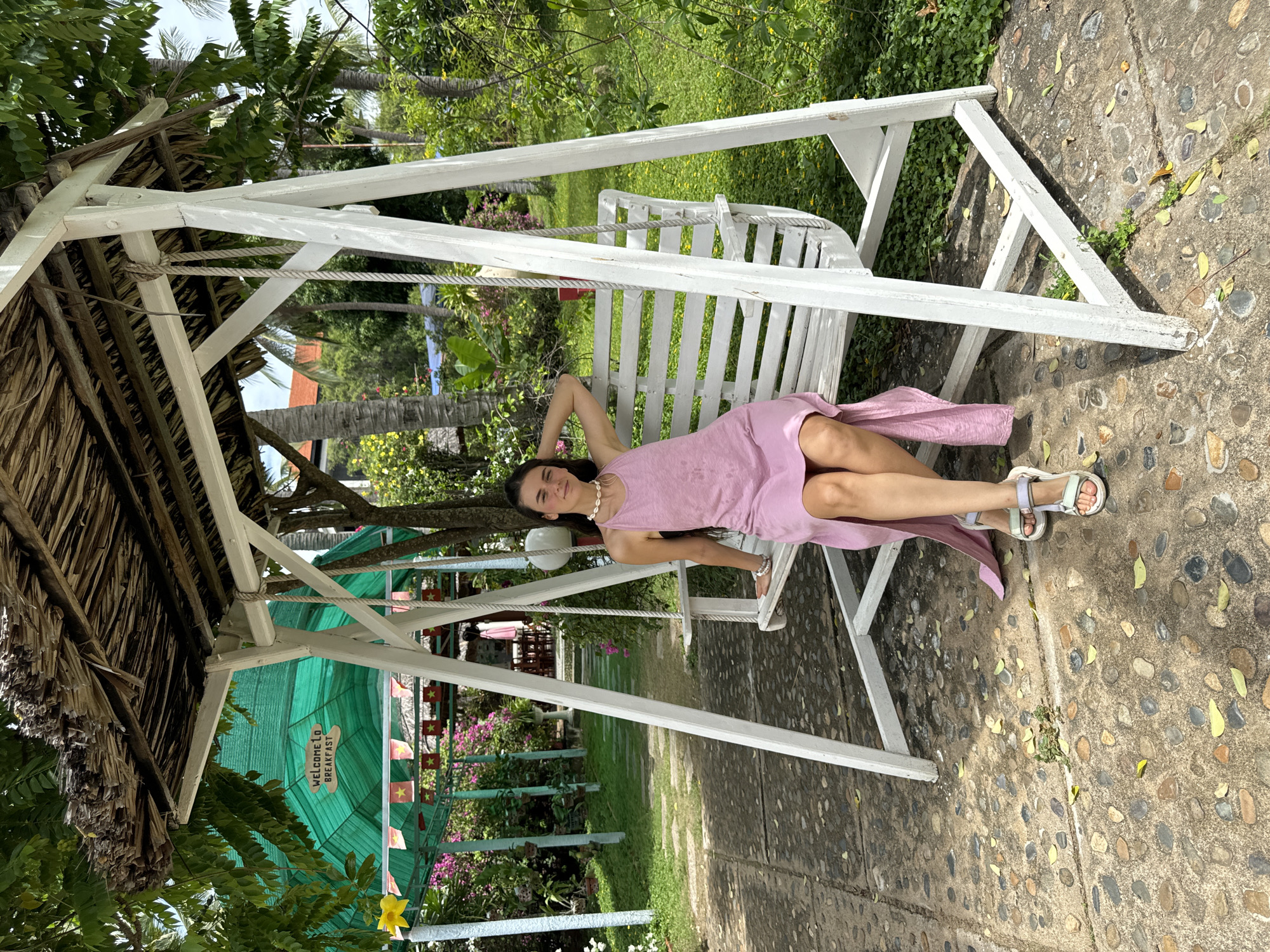 A woman is depicted relaxing on a white wooden swing outdoors in a bright, tropical setting. She lies on her back on the swing, with her head propped by one arm and the other arm resting on her lap. She wears a light pink sleeveless dress, a white shell necklace, and light-colored sandals. Her dark hair is loose, and she looks towards the camera with a gentle smile.
The swing is a white slatted bench, suspended by ropes from a white wooden A-frame structure. The ground beneath is a paved path made of embedded pebbles, with scattered green and yellow leaves.
The surrounding environment is lush and green, featuring various trees and plants, including palm trees. To the left, a traditional thatched roof structure is visible. In the background, a large green shade net or awning is strung with small flags that have a red background and a yellow star, resembling the flag of Vietnam. A wooden sign, shaped like a footprint, is attached to a post beneath the awning, bearing the white text "WELCOME TO BREAKFAST." The scene is well-lit by bright daylight, casting distinct shadows, indicating a sunny day.