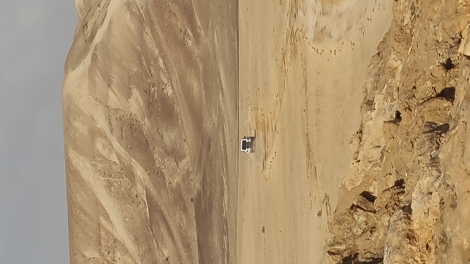 A white car is driving on a dirt road in the desert. The scene is likely located in the Tamar Regional Council, Israel, based on visual characteristics of the landscape. The environment includes desert terrain, a dirt road, and possibly rock formations or cliffs. There are no visible people or other cars in the frame. The action is the car moving along the road.