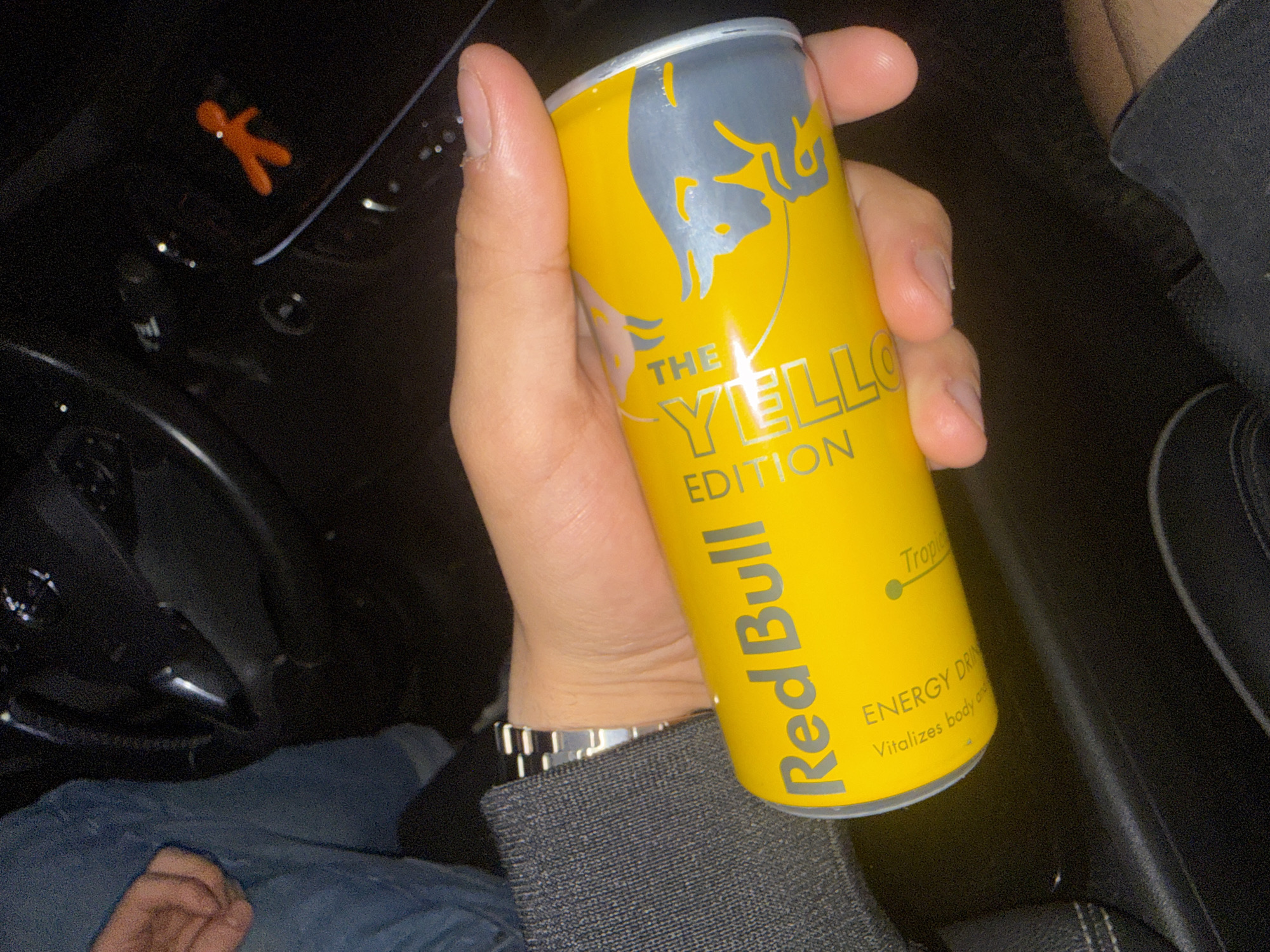 The image captures a close-up, dimly lit scene within the interior of a vehicle. In the foreground and center, a light-skinned hand holds a yellow Red Bull "The Yellow Edition" energy drink can. The can features the Red Bull logo in silver and two gray bull silhouettes, with "ENERGY DRINK" and "Tropical" (partially visible) printed on it. The hand is wearing a metallic, multi-link watch on its left wrist, with a dark, ribbed fabric sleeve visible around the wrist area.
To the left of the central hand, a second person's left hand and a portion of their blue denim pants are visible, suggesting they are seated. In the upper left background, part of a dark steering wheel and dashboard elements are discernible. On the right side of the dashboard, a small, orange, stylized human-like figure is attached, possibly an air freshener or decorative item. The overall lighting is low, with some reflections visible on the dark surfaces of the car interior. No specific landmarks or unique environmental features are present to confirm the city of Varna, Bulgaria, as the location.