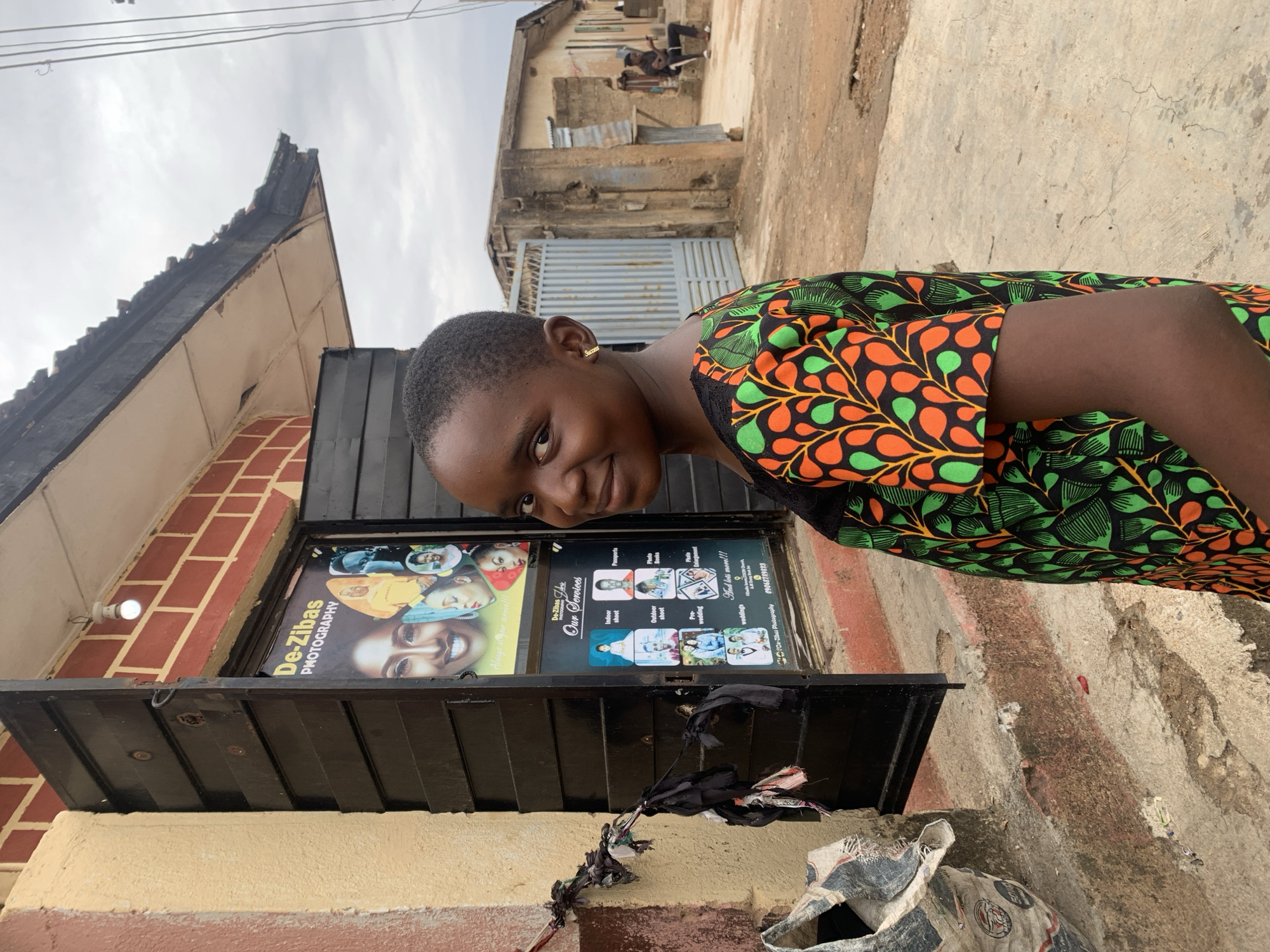 A young person, likely a girl or young woman, smiles warmly at the camera in an outdoor setting in Jos, Nigeria. She has short, dark hair and wears a vibrant, patterned top with green, orange, and black leaf-like motifs. A small gold earring inscribed with "Success" is visible in her left ear.
She is standing in front of a building that houses "De-Zibas Photography," as indicated by a prominent sign. The sign details various services offered, including "Passports," "Indoor shoot," "Outdoor shoot," "Photo Books," "Pre-wedding," and "weddings," along with a contact number, "0906879823." A light bulb is mounted on the wall near the sign. Below the sign, a dark awning or shutter is visible, from which some dark fabric items are hanging.
In the background, under an overcast sky, typical urban structures like other buildings with weathered facades, corrugated metal gates, and roofs are discernible, suggesting a street or neighborhood environment. The ground appears to be a mix of concrete and dirt. The overall impression is a cheerful, candid moment captured outside a local photography business during daylight hours.