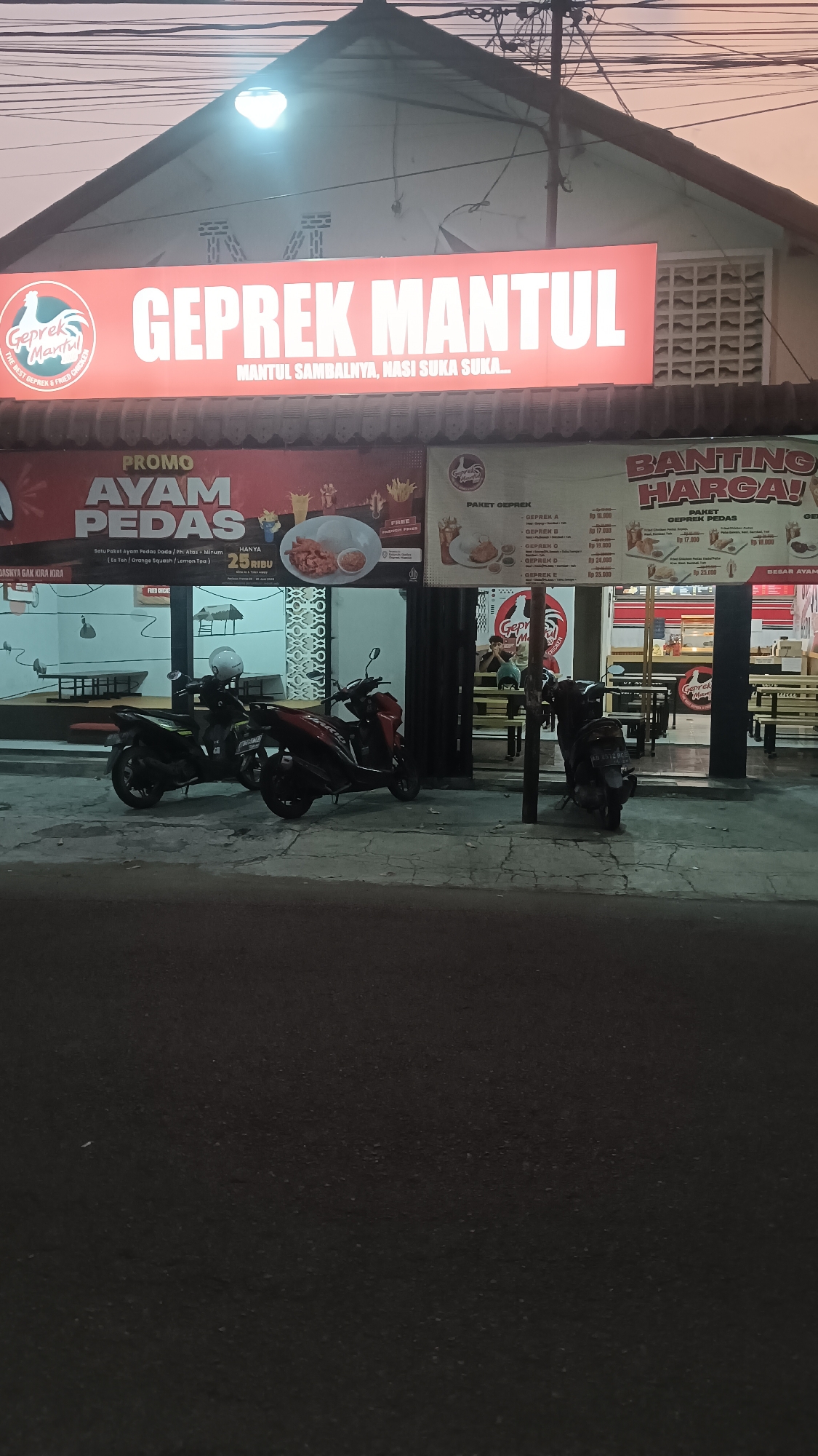 The image depicts the exterior of a "Geprek Mantul" restaurant located in Sragen Tengah, Indonesia. The restaurant features a large, illuminated sign with the name "GEPREK MANTUL" in prominent lettering. Below the main sign, a smaller sign reads "MANTUL SAMBALNYA, NASI SUKA SUKA...", indicating the restaurant's specialty dishes.
Several promotional banners are visible. One banner advertises "PROMO AYAM PEDAS" (Spicy Chicken Promo) with the phrase "HANYA 25 RIBU" (Only 25 Thousand Rupiah) and details about the package including "Satu Paket Ayam Pedas Dada / Fillet + Es Teh / Orange Squash / Lemon Tea" (One Spicy Chicken Breast / Fillet Package + Iced Tea / Orange Squash / Lemon Tea). Another banner on the right side of the image announces "BANTING HARGA!" (Price Crash!) with "PAKET GEPREK PEDAS" (Spicy Geprek Package).
In front of the restaurant, two motorcycles are parked. One is a black motorcycle and the other is a red motorcycle. Beyond the motorcycles, the interior of the restaurant is partially visible, showing tables and chairs, some of which appear to be wooden benches and tables. The restaurant entrance is open, and some customers are seated inside. The overall environment suggests a casual dining establishment. The time of day appears to be late afternoon or early evening, given the lighting conditions.