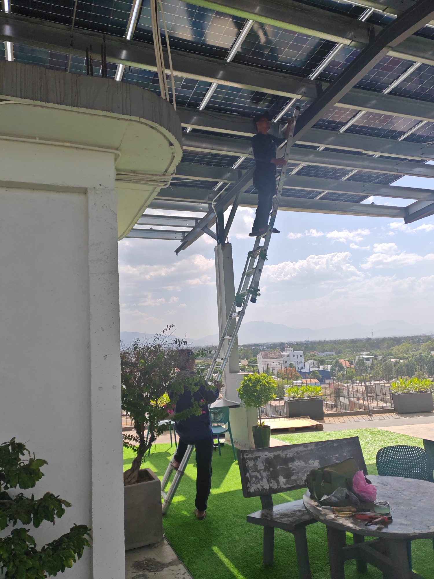 On a rooftop in Đà Nẵng, Vietnam, two individuals are installing solar panels. One person stands on an aluminum ladder beneath a metal frame supporting the solar array, appearing to be making adjustments. The other person is positioned on a lower section of the same ladder, also engaged with the structure. The rooftop is covered with artificial turf, and potted plants, including a small tree, are visible. In the background, a cityscape with buildings and distant mountains under a cloudy sky can be seen. A concrete table with chairs and some tools are also present on the rooftop.