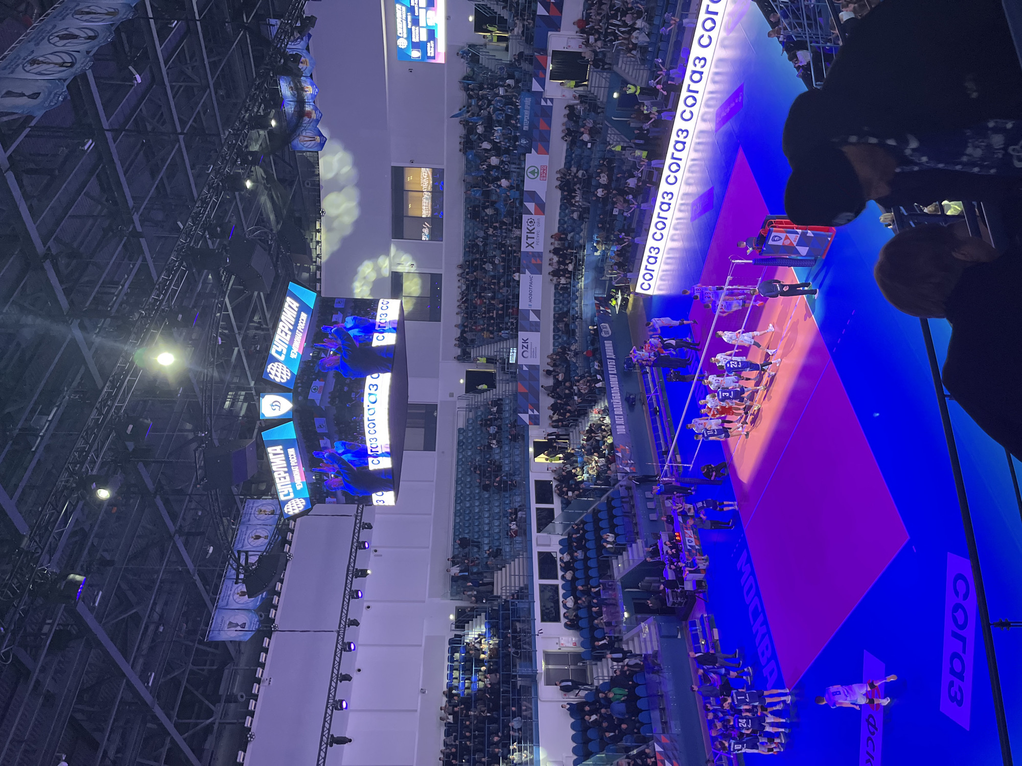 The image shows a volleyball game taking place in a large indoor arena in Moscow, Russia. The court is colored in blue and pink. There are two teams lined up on either side of the net, wearing white and blue uniforms. Spectators fill the stands surrounding the court. There are advertisements for various Russian companies displayed, including "СОГАЗ", "OZK", "XTKO", and "HOBOTРАНС". Banners hanging from the ceiling display the words "СУПЕРЛИГА ЧЕМПИОНАТ РОССИИ", indicating that the game is part of the Russian Super League championship.