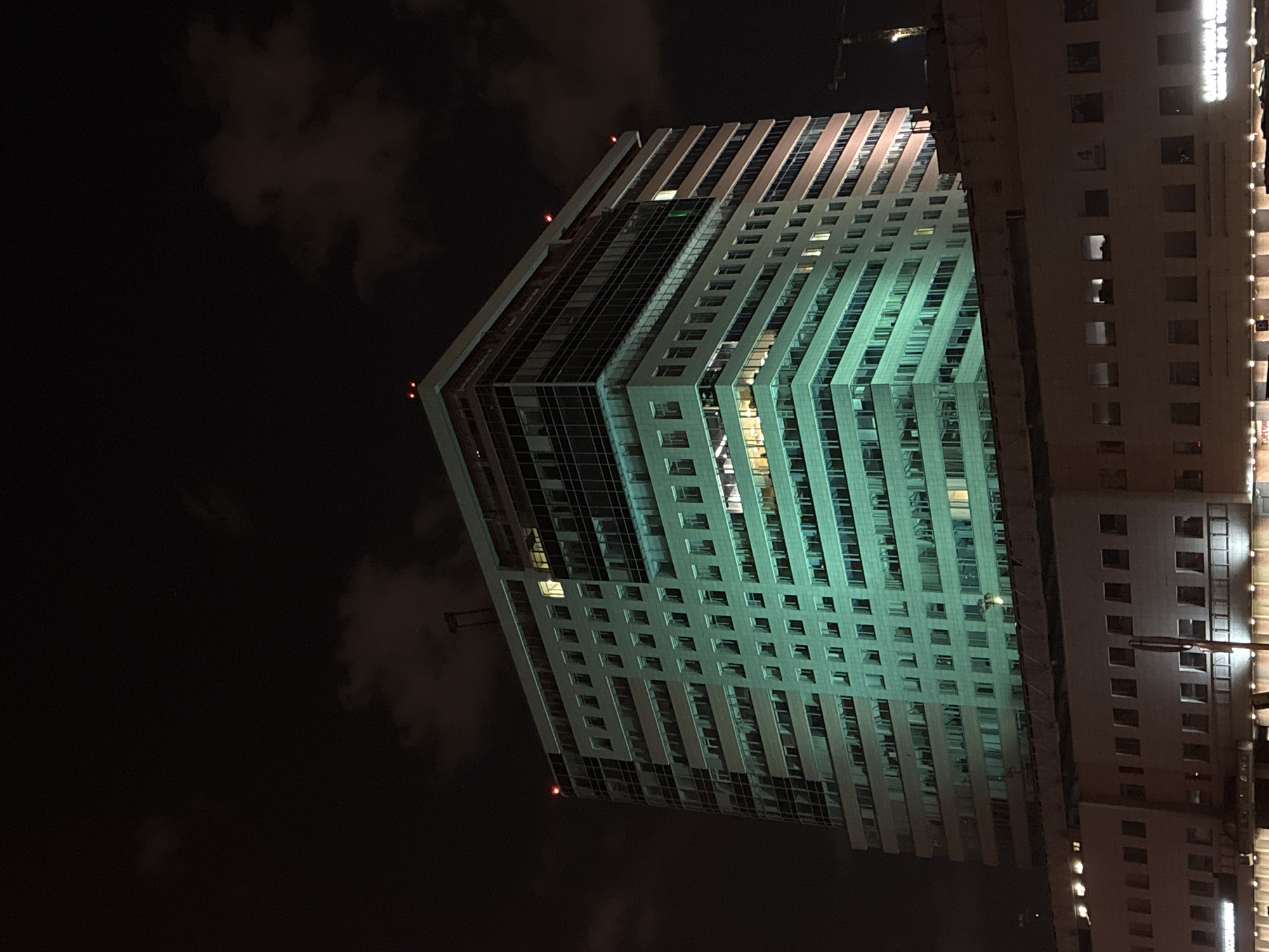 The image captures a night scene in Moscow, Russia, featuring illuminated buildings. The primary structure is a modern, high-rise building characterized by a grid-like facade of windows. A light source illuminates the building. Adjacent to the high-rise is another building with a similar window pattern. The sky is dark with some cloud cover.