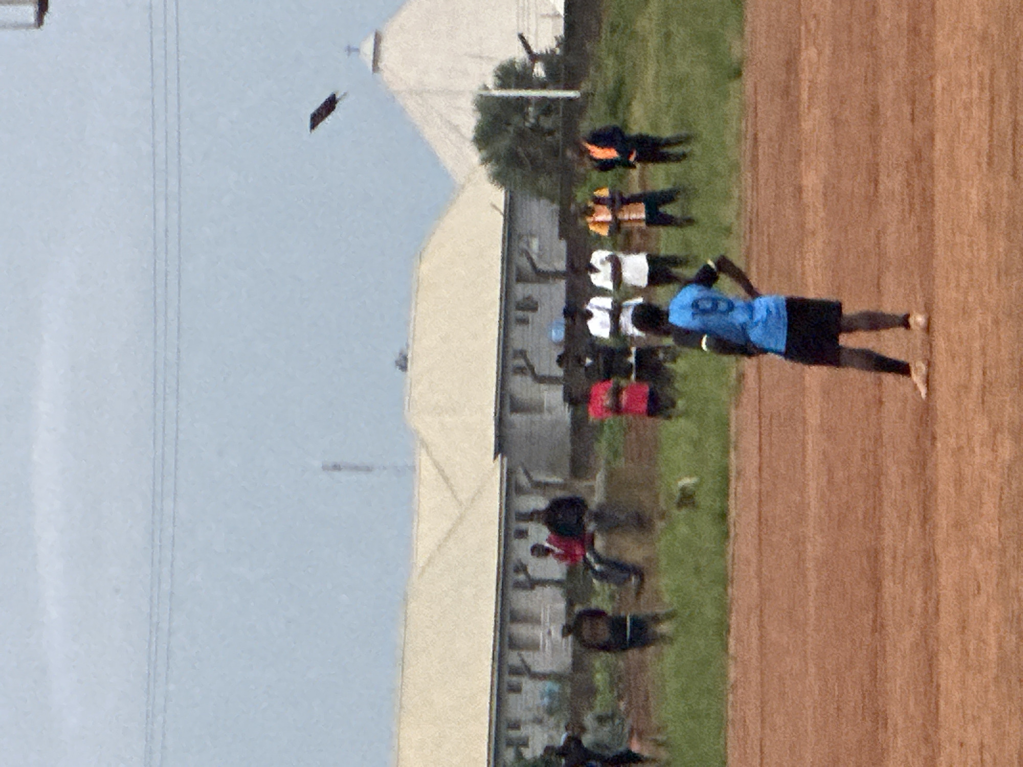 The image captures a scene on an outdoor dirt field, likely a sports ground, in Du, Nigeria. In the foreground, a person in a blue jersey with the number 19 and black shorts stands facing away from the camera, with one hand on their hip. The field itself is reddish-brown dirt. In the midground, a group of individuals are visible, some standing and others seated on a low wall or ledge. One person wears a red shirt, another a black shirt, and some are in white shirts. Behind them are buildings with light-colored, pitched roofs. Further in the background, a large, white, domed structure, possibly a church, is visible, along with trees and utility poles. In the upper portion of the image, power lines stretch across a cloudy, pale blue sky. A dark, elongated object, possibly a kite or a piece of debris, is airborne in the sky, to the left of the domed building. The overall atmosphere appears overcast.