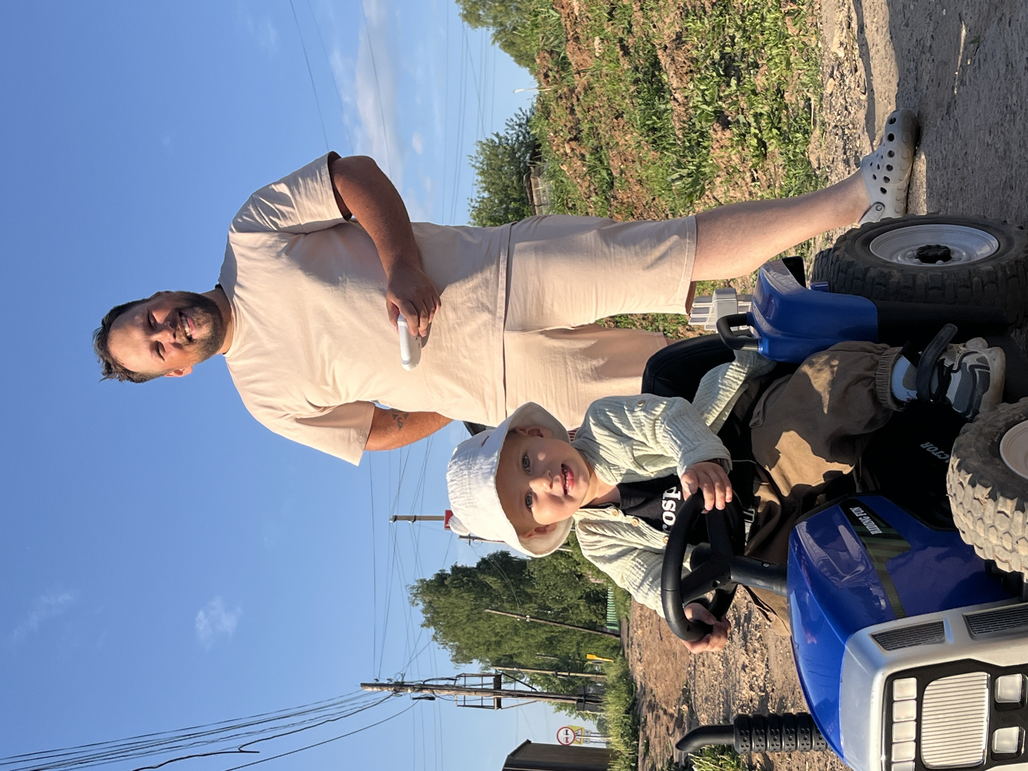 A man and a child are on a toy tractor in Фоки, Russia. The man, with short dark hair and a beard, is wearing a light brown t-shirt and shorts, and light grey Crocs, is standing on the back of the tractor. He has a white object in his right hand. The child, wearing a white hat, a light green sweater over a dark t-shirt, is sitting in the driver's seat, holding the steering wheel. The tractor is blue and white. The background includes a dirt road, a grassy area, power lines, trees, and a clear blue sky. A speed limit sign is visible behind the tractor.