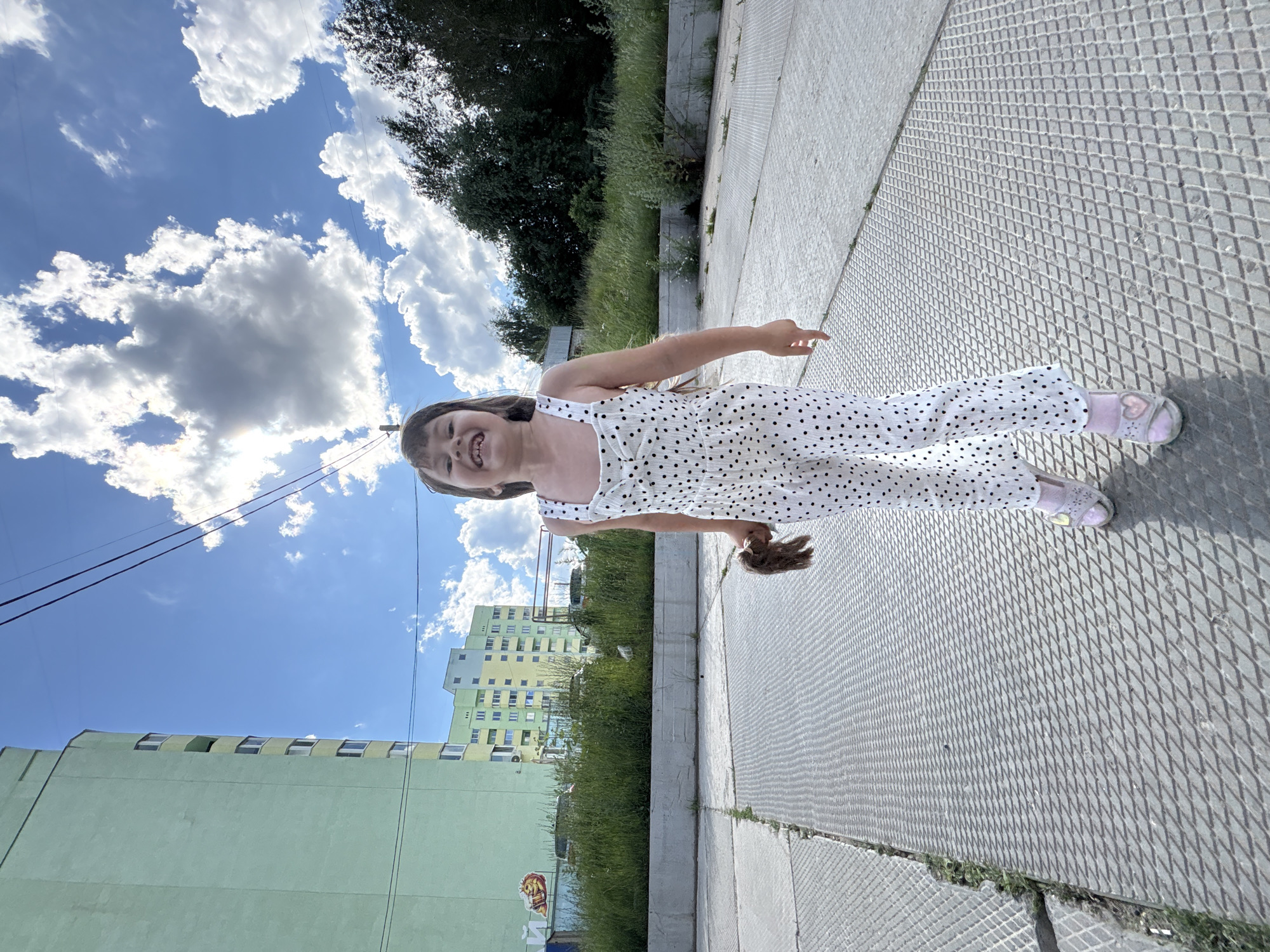 A young girl with dark hair and a wide smile stands in the foreground of the image. She is wearing a white polka dot jumpsuit and pink sandals. In her right hand, she holds a brown stuffed toy. The girl is positioned on a concrete surface with a diamond-patterned texture, likely a sidewalk or plaza. Behind her, a line of green grass separates the concrete from a low concrete wall. Further back, a group of tall trees forms a dense line. In the distance, several apartment buildings are visible, with one prominent light green building on the left and a multi-story yellow and green building to its right. The sky is bright blue with scattered white clouds, and power lines are visible stretching across the upper portion of the frame. The scene appears to be an urban outdoor environment.