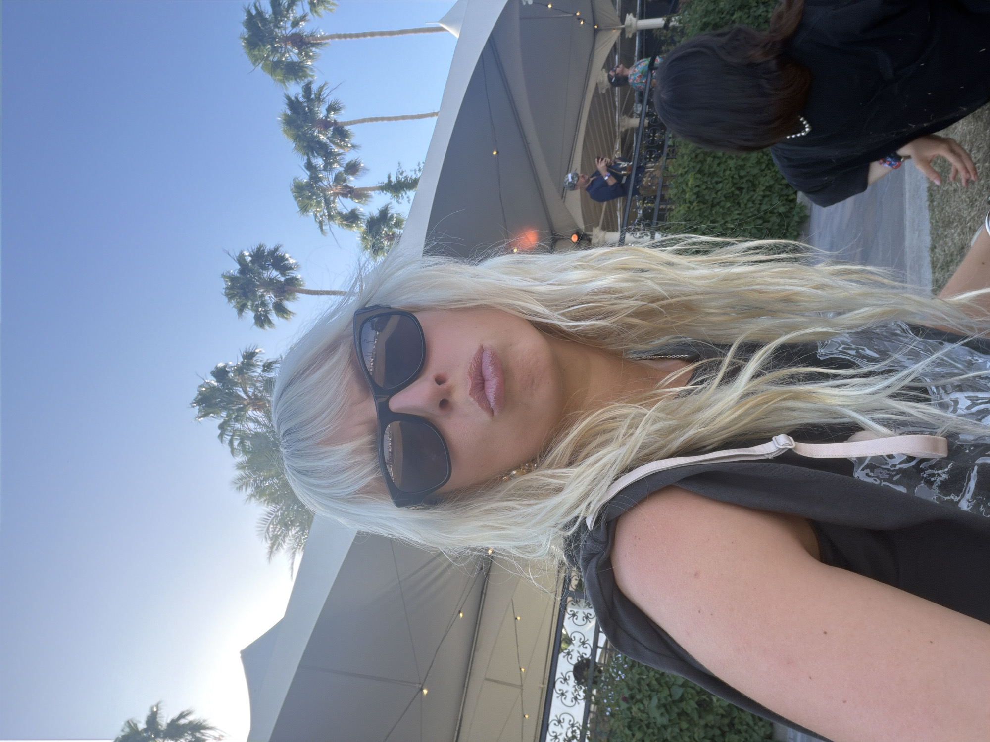 The image shows a selfie of a fair-skinned woman with blonde hair and dark sunglasses, taken outdoors. She is wearing a dark top with a lighter-colored strap visible. Palm trees are visible against a bright blue sky. A tent-like structure is in the background, with a metal fence and some people faintly visible behind it. The location is likely in the city of Indio, California, United States of America, based on the presence of palm trees and the style of the outdoor structure.