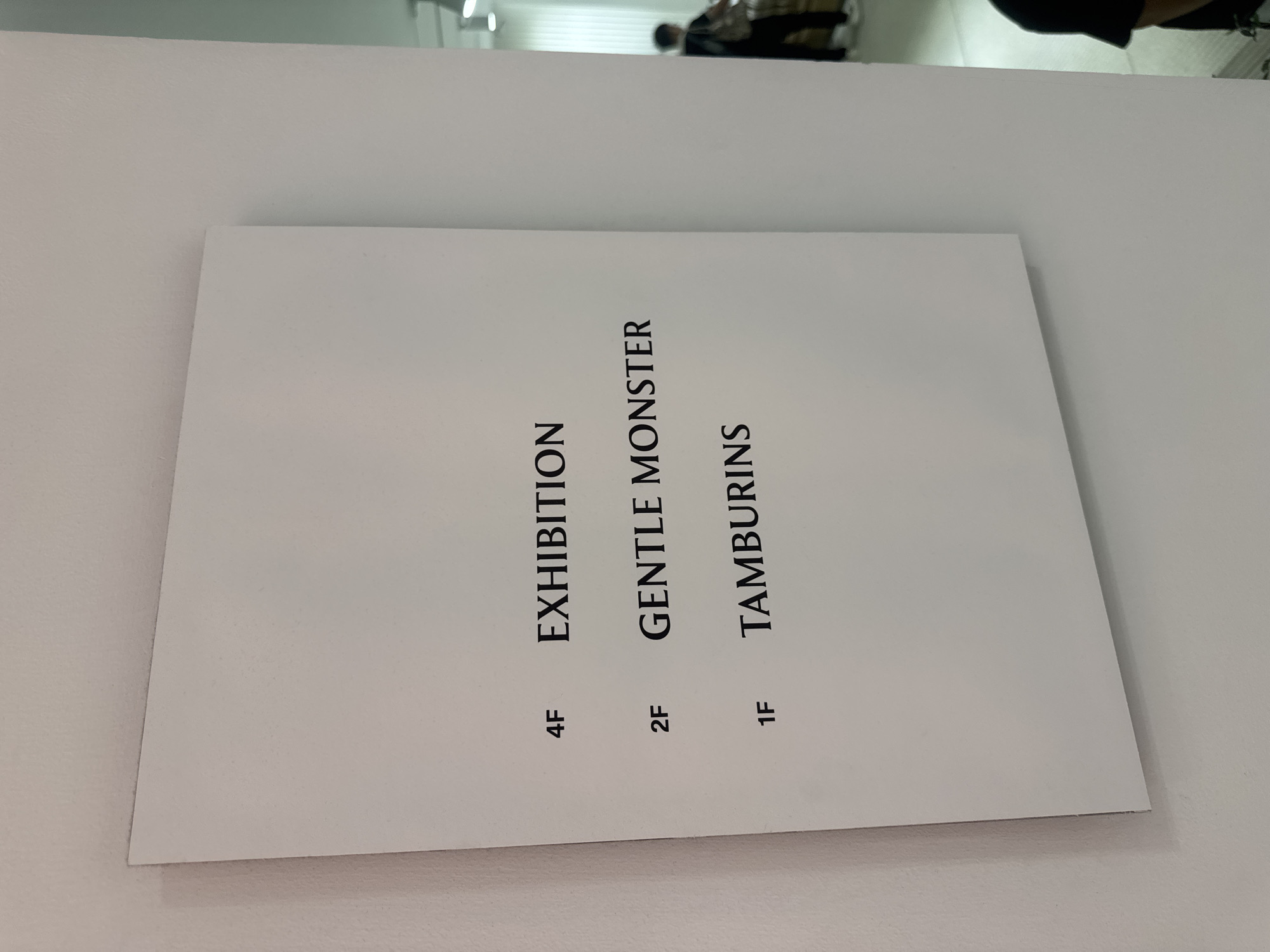 The image shows a white sign mounted on a white wall. The sign lists "4F EXHIBITION", "2F GENTLE MONSTER", and "1F TAMBURINS". A person with dark hair, wearing a dark t-shirt and dark pants, is visible in the blurred background on the right side of the image. Another person is partially visible in the lower right corner. The environment appears to be an indoor space with a bright ceiling light. The sign is positioned in the foreground and appears to be at eye level. The wall texture is visible to the left of the sign. The overall lighting is bright.