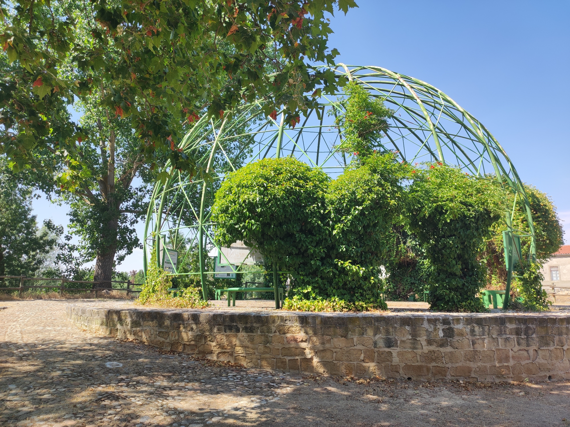 The photograph captures an outdoor scene in the city of Huerta, Spain. In the foreground, a stone wall borders a cobblestone pathway. Beyond the wall, a large, green, domed structure, likely a trellis or arbor, is covered with lush green foliage and some red flowers. Several green benches are visible beneath the structure. Large trees with green leaves and some with red-tinged leaves are present in the background, along with a wooden fence and a building with a window. The sky is clear and blue.