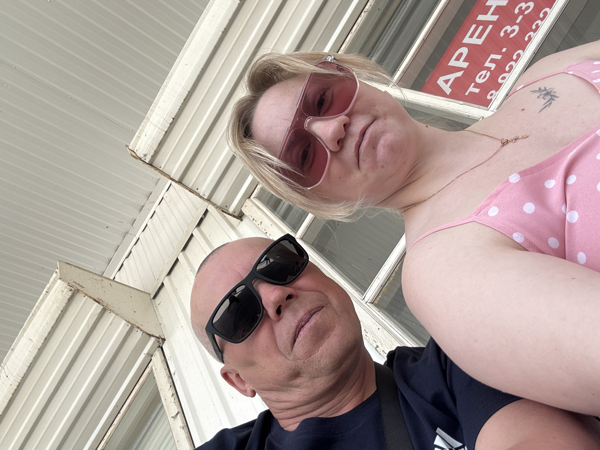 Two people, a man and a woman, are in the frame. The man, bald, is wearing black sunglasses and a dark shirt. The woman has blonde hair, wears pink sunglasses, and a pink polka-dot dress. She has a small tattoo on her upper chest and wears a gold necklace. They are standing in front of a building with metal siding and windows. A sign with Cyrillic text is visible in the window, which includes the word "АРЕНД" and a phone number. The image was taken in the city of Chaikovsky, Russia.