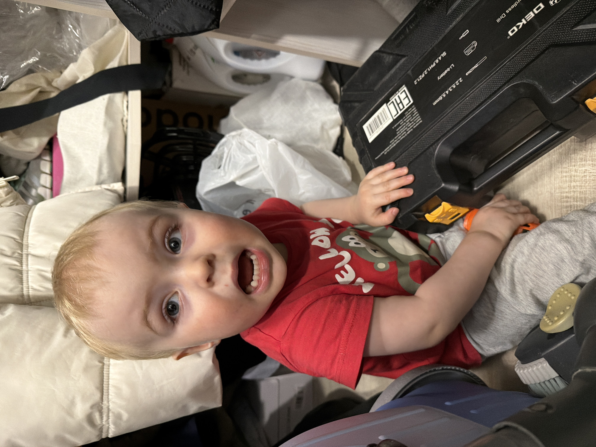 A child with blonde hair and light skin is lying on the floor, next to a black DEKO cordless drill case. The child is wearing a red shirt with a cartoon bear and grey pants. The child's mouth is open and their eyes are wide, as if they are surprised or excited. The background is cluttered with a variety of items, including what appears to be clothing, plastic bags, and other objects. The text "MADE IN" and "EAC" is visible on the drill case. There is no discernible information about the city of Votkinsk, Russia.