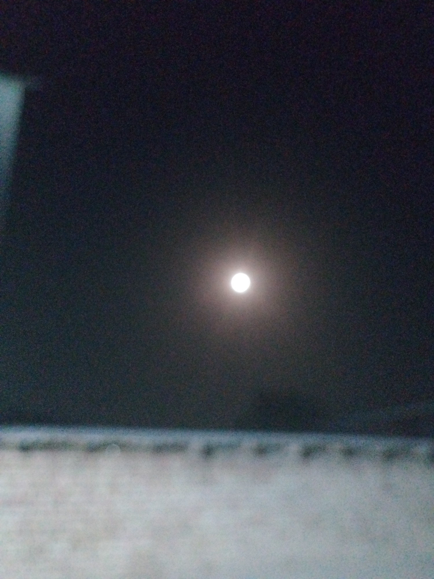 The image depicts a night scene with a full moon prominently featured in the dark sky. The moon is a bright, circular object, slightly diffused by the atmosphere. The sky is a deep, dark gray, suggesting it is nighttime. In the foreground, there is a blurry, horizontal band of lighter color, possibly indicating a surface like a building or ground, but it is out of focus and not clearly defined. There are no people or any discernible activity visible in the scene. The image appears to have been taken from a low vantage point. The location is identified as Социализм, Kyrgyzstan.