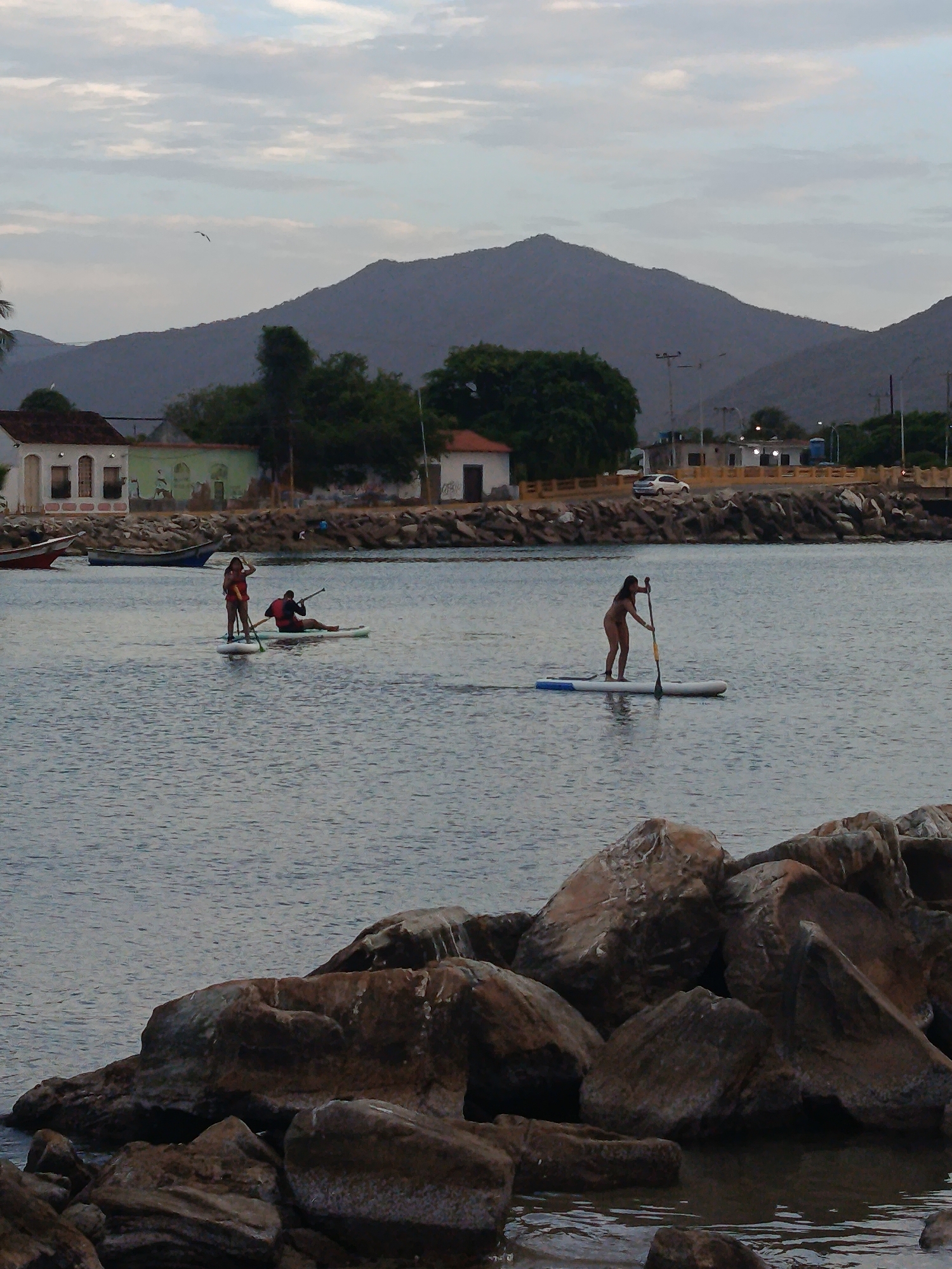 This image depicts a view of the bay in Juan Griego, Venezuela, with a calm sea and a mountainous backdrop. In the foreground, large rocks form a breakwater. The water is populated by three individuals on paddleboards. On the left, two people are on a single paddleboard; one is standing and the other is seated. Further to the right, a lone person is paddleboarding. In the middle ground, a coastline features several buildings, trees, and a road with a white car. The sky above is overcast with scattered clouds.
