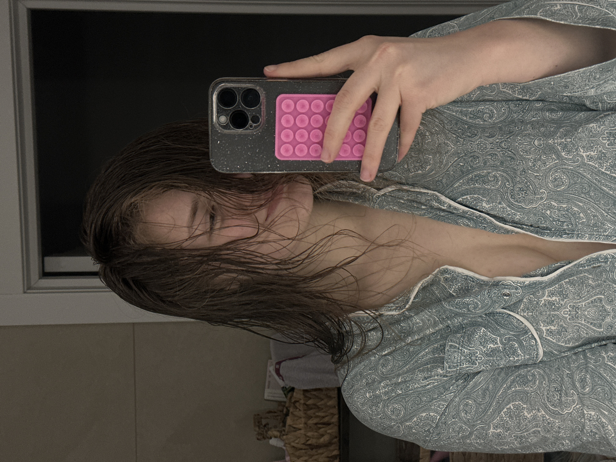 A person is taking a selfie in a bathroom, holding a cell phone in their right hand. The phone has a grey speckled case and a pink grip pad. The person has wet brown hair and is wearing a blue paisley patterned pajama top with white piping. The background includes a window with a black pane, a tiled wall, and a laundry basket with a towel. No evidence indicates the location is Kazan, Russia.