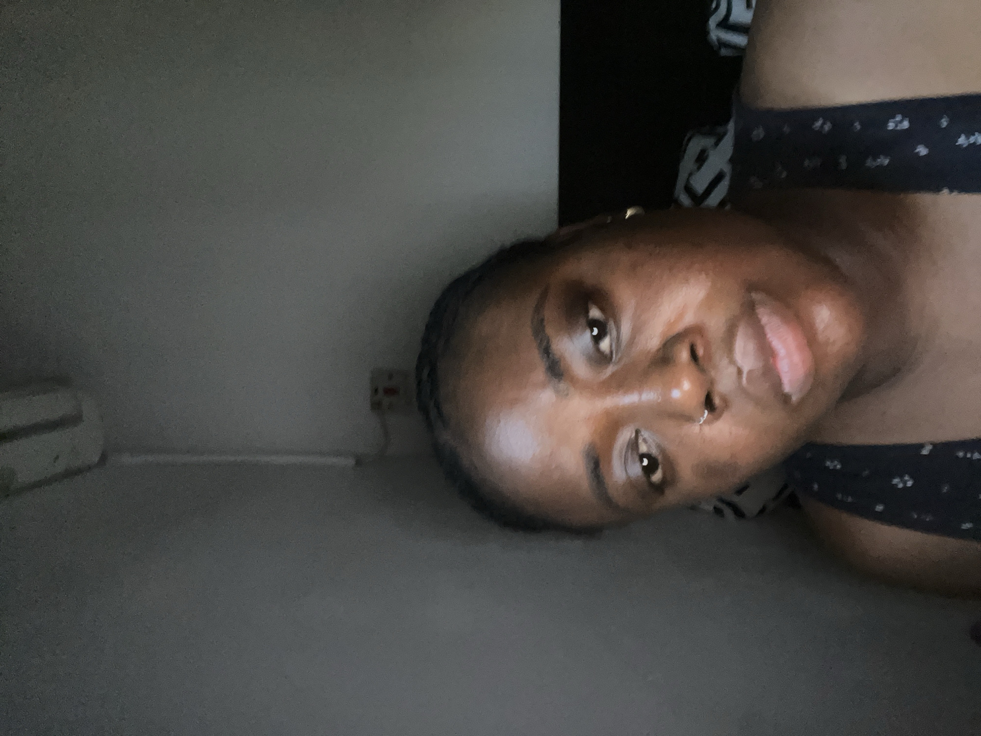 A selfie taken indoors of a person with dark skin and short, dark hair. The person is wearing a dark tank top with a small white pattern. The background includes a light grey wall, an electrical outlet, a white air conditioning unit, and a dark piece of furniture. The location is potentially a private residence. Location in the city of Abuja, Nigeria cannot be verified.