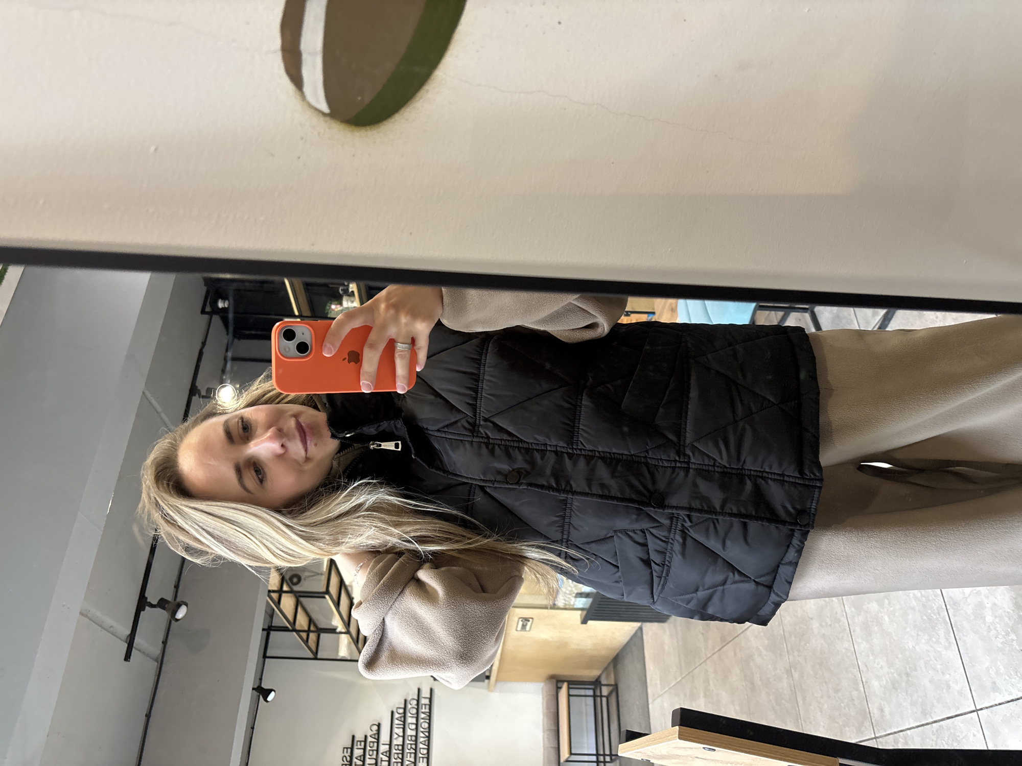 A young adult female is captured in a mirror selfie within a modern establishment in Chaikovsky, Russia. She stands centered in the reflection, holding an orange iPhone in her right hand, which partially obscures her lower face and right shoulder as she looks down at the phone's screen. Her long, light blonde hair, parted slightly off-center, cascades over her right shoulder. She wears a black quilted puffer vest with a stand collar and snap closures over a zipper, layered over a light beige fleece long-sleeve top with a high collar and sleeves pushed up on her right arm. Matching light beige wide-leg trousers complete her casual ensemble. A silver-toned ring is visible on her right ring finger, and a delicate chain bracelet adorns her right wrist.
The reflected background reveals an interior space, consistent with a contemporary cafe. The ceiling is exposed grey concrete with black track lighting fixtures housing multiple spotlights. The light-colored walls feature three-dimensional black lettering displaying menu items in both English (e.g., 'DAILY BREAD,' 'COLD BREW,' 'LEMONADE') and Cyrillic text. To the reflected left, a light wooden bench with an upholstered seat and a small wooden table with black metal legs are present. Further back, an industrial-style shelving unit with wooden shelves and black metal frames is visible. The floor is covered with large, light grey rectangular tiles. To the reflected right, a partial view of a soft blue upholstered seating area and a circular wall decoration, half green and half brown with a white horizontal stripe, are visible. The mirror itself is framed by a thin black border, and the adjacent wall outside the mirror is plain off-white with minor imperfections.