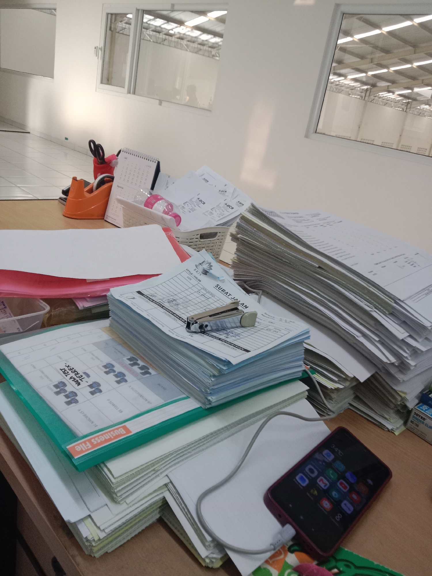 The image depicts a cluttered desk in what appears to be an office environment. Several stacks of papers, some with writing and labels, are scattered across the surface. Office supplies such as a stapler, a tape dispenser, and a pair of scissors are visible among the documents. A desk calendar is also present. In the foreground, a smartphone is lying on the desk, plugged into a charger. The background shows a brightly lit interior with windows that offer a view of a large industrial or warehouse-like space with a metal roof structure. The flooring in the background appears to be tiled. The overall impression is of a working office with ongoing administrative tasks. No specific details about the city of Semarang, Indonesia, can be inferred from the image itself.