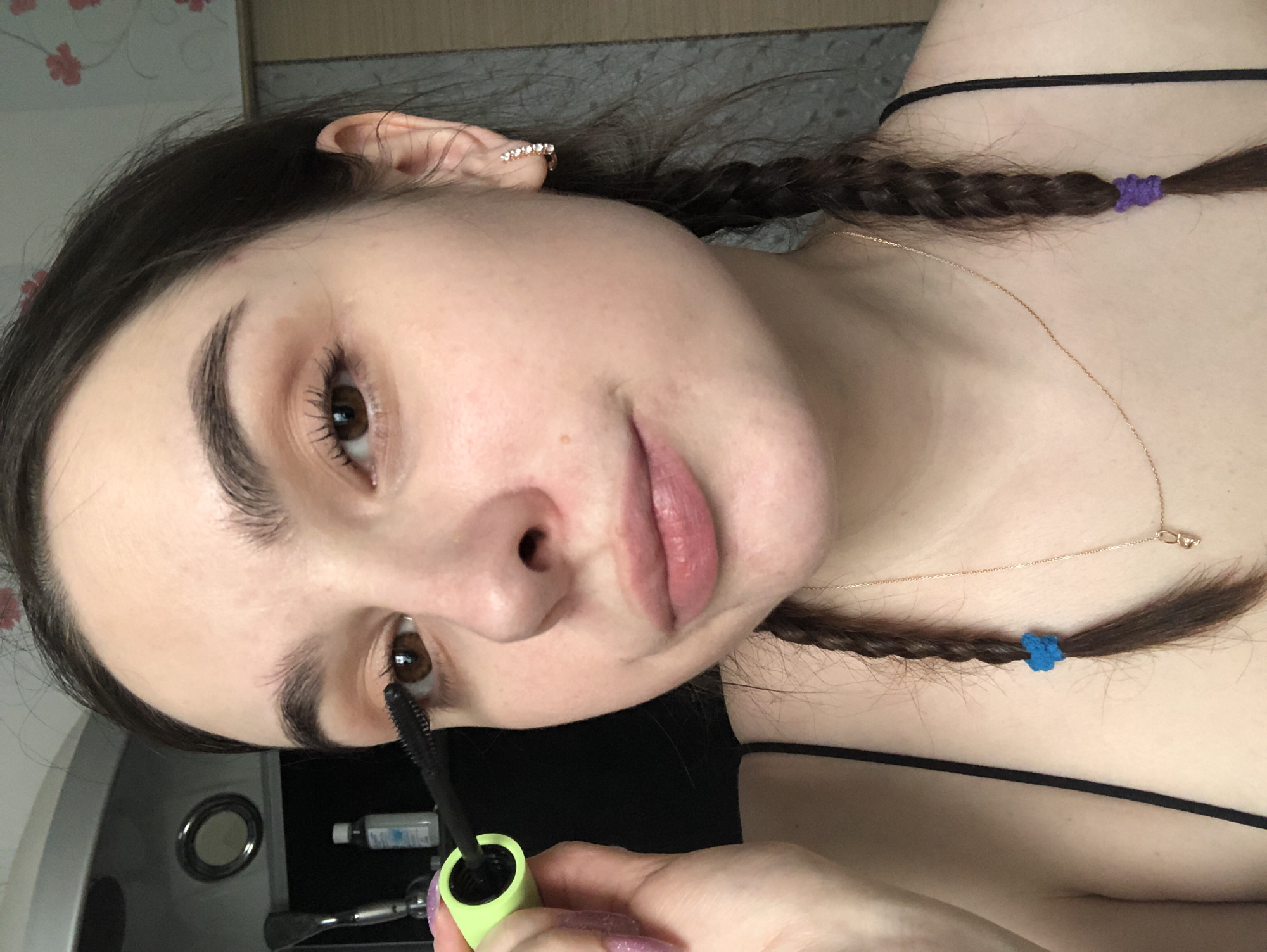The scene is an indoor, close-up view of a young adult female applying mascara to her right eye. Her fair complexion, brown eyes, and dark brown hair, styled into two braids with blue and purple elastic ties, are prominent. Her left hand, featuring long, purple glitter nail extensions, holds a neon green mascara wand with a black brush. She wears a black spaghetti strap top, a gold-colored hoop earring with clear stones in her right ear, and a thin gold-colored necklace with a small pendant. The background on the left includes a dark counter or shelf with a small white bottle and a circular mirror. On the right, wallpaper with a red floral pattern is visible, alongside a light-colored wooden door or cabinet. The image contains no visual information to establish its location in the city of Noviy, Russia, as requested.