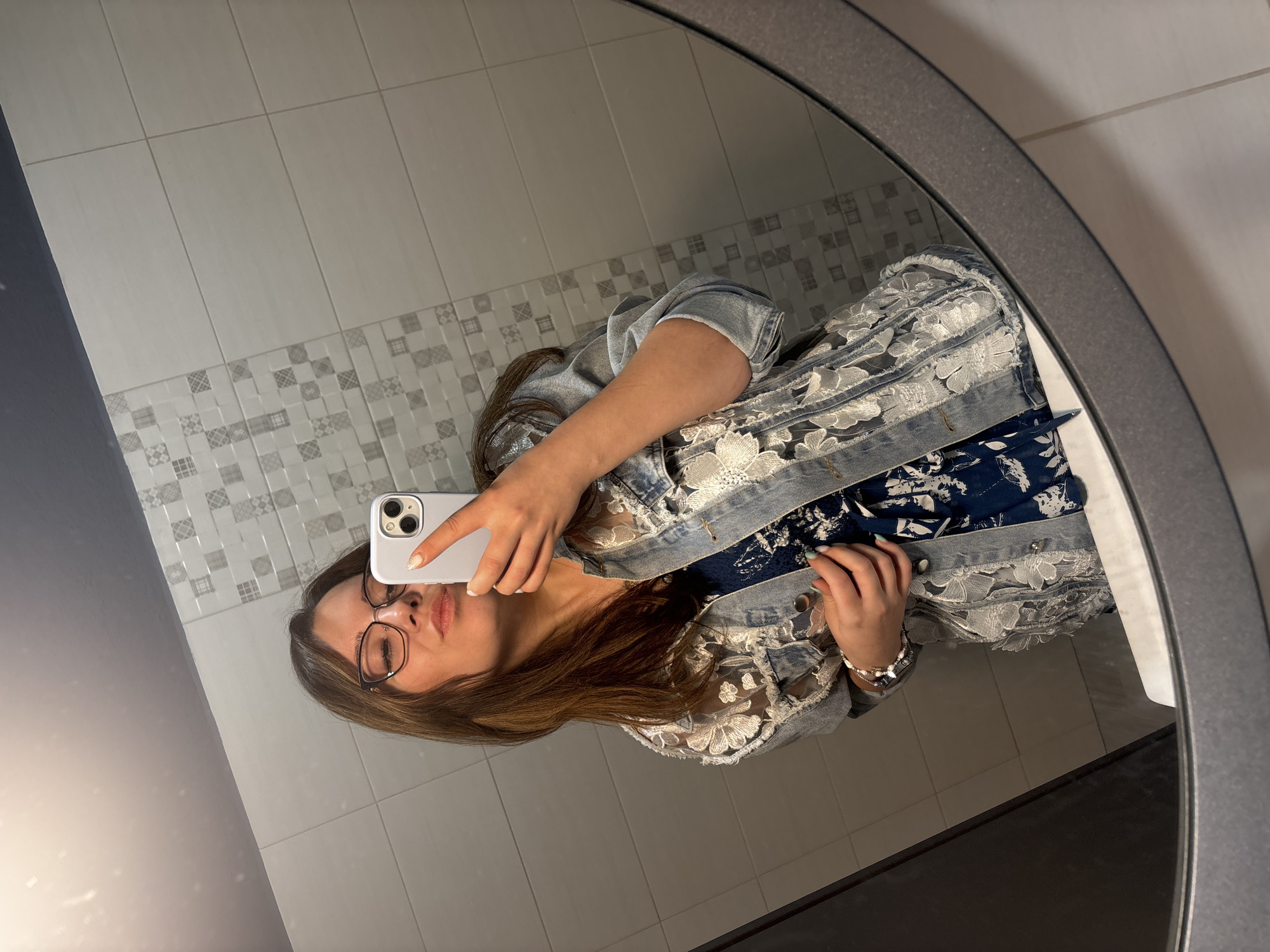 A young woman with brown hair and glasses is taking a mirror selfie in a bathroom. She is wearing a blue floral dress and a denim jacket with lace details. She is puckering her lips for the photo and holding her phone up to take the picture. The bathroom has tiled walls and a large oval mirror.