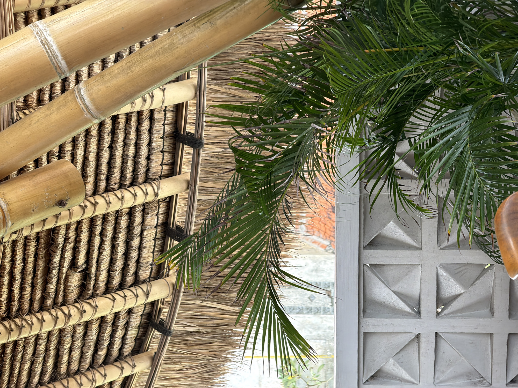 This is a medium shot, eye-level, outdoor photograph taken in Kuta Selatan, Indonesia, showing a tropical-themed interior or outdoor seating area. The foreground is dominated by a lush green palm plant, with its fronds obscuring part of the background. The plant is positioned in front of a textured, grey, concrete block wall featuring a repeating pattern of raised squares with triangular indentations. Behind the wall and palm, a light-colored structure with terracotta roof tiles is partially visible.
The ceiling is constructed of natural materials: bamboo poles form the structural beams, with tightly woven dried grass or thatch forming the roof covering. Dark rope is used to bind the bamboo elements. The lighting suggests it is daytime, with natural light filtering through the structure, creating a warm, tropical ambiance. There are no people or animals visible in the image. The overall impression is one of a relaxed, natural, and inviting space.
