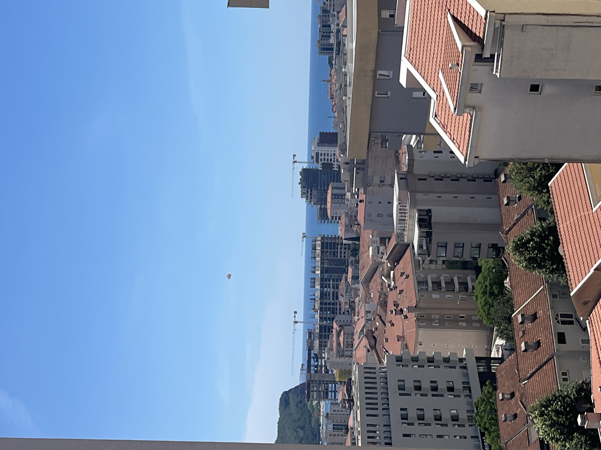 The image, best viewed horizontally, captures a vibrant coastal cityscape under a clear, bright blue sky on a sunny day in Budva, Montenegro.
In the foreground and midground, a dense urban environment unfolds. Numerous multi-story buildings with distinctive red-tiled roofs are prominent, showcasing a traditional Mediterranean architectural style. Alongside these, more modern, taller buildings are visible, some of which appear to be under construction, with several tower cranes dotting the skyline. Green foliage from trees is interspersed among the buildings, adding a touch of nature to the urban setting.
Beyond the city, a calm, blue body of water, likely the Adriatic Sea, stretches towards the horizon. In the far distance, a verdant, tree-covered mountain rises from the coastline.
The sky is a vast and cloudless blue. A small, red and white paraglider is a notable subject, suspended high in the sky, indicating recreational activity. The bright lighting confirms it is daytime. No discernible text is visible in the image.