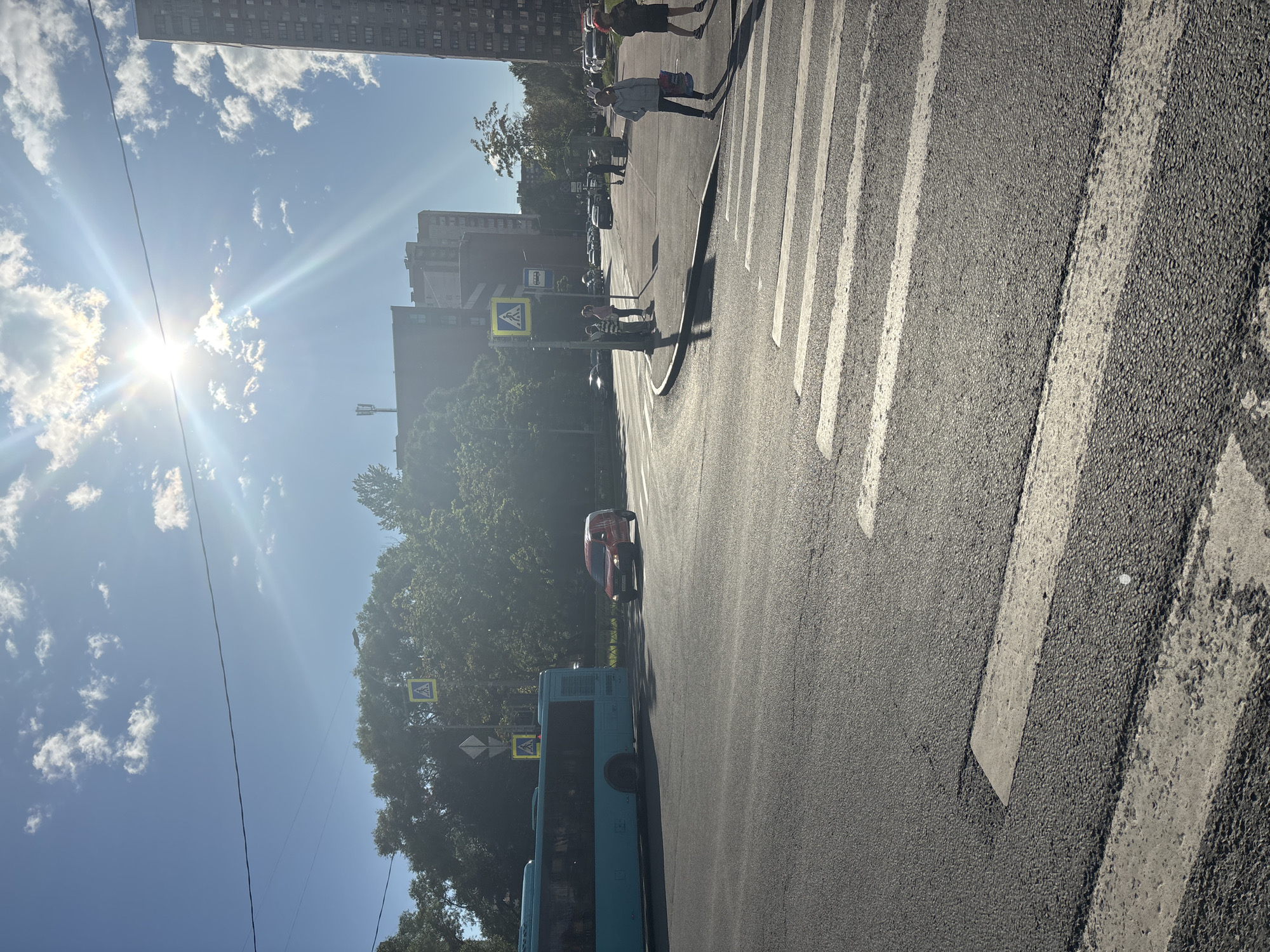 The image depicts a sun-drenched street scene in an urban environment, possibly in Russia, with a prominent crosswalk in the foreground. The sky is a bright blue, with scattered white clouds and the sun shining intensely, casting lens flares. A light blue bus is visible on the left, partially obscuring a red van stopped at what appears to be an intersection. Several pedestrians are present, some waiting at a crosswalk and others walking across. Yellow pedestrian crossing signs are mounted on poles at the intersection. Tall apartment buildings and trees line the background. The asphalt road shows wear and tear, with white painted lines marking the crosswalk. A wire stretches across the upper portion of the frame.