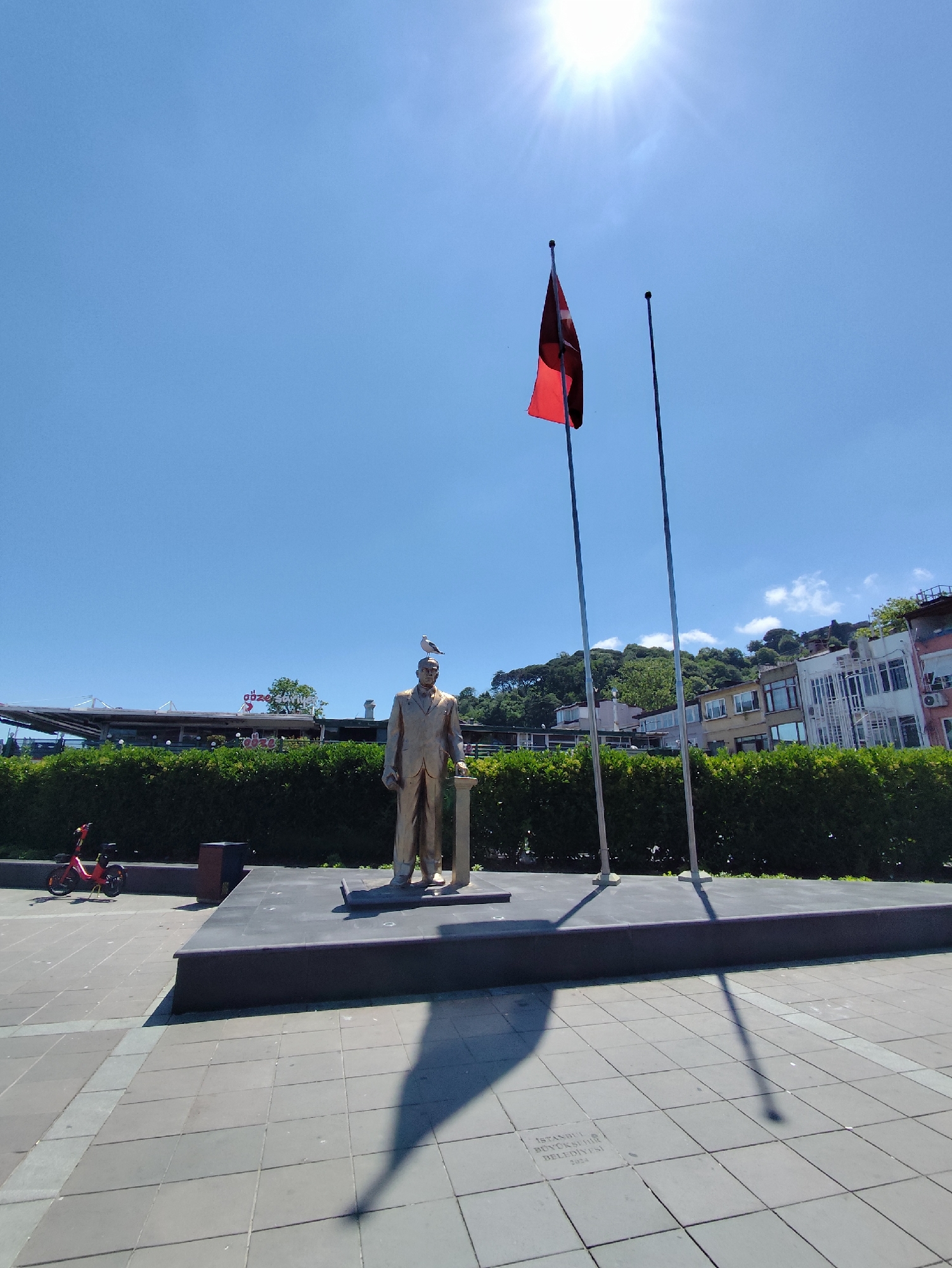 This is a full-shot, eye-level, outdoor, daytime, medium-angle photograph of a statue of a man in a suit. The statue is made of bronze and stands on a dark pedestal. A bird is perched on the statue's head. Two flagpoles stand behind the statue, one flying a Turkish flag. In the background, there is a dense green hedge, a building with awnings, and further back, a hillside with trees and buildings. The sky is clear and bright blue with the sun shining intensely, creating long shadows on the paved ground in the foreground. To the left of the statue, there is a red electric scooter. The paving stones in the foreground have some visible text etched into them.