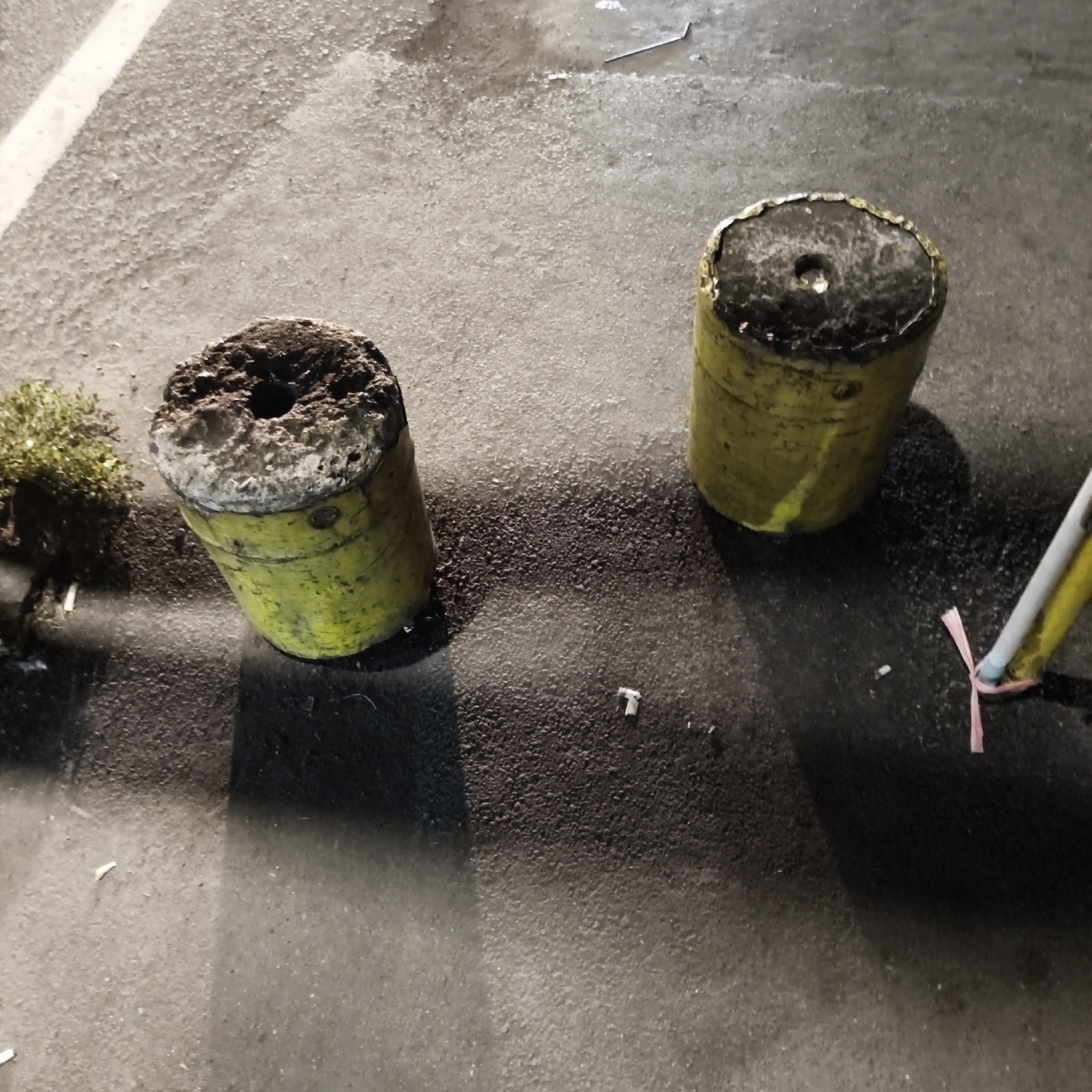 The image depicts two cylindrical, yellow concrete bollards on an asphalt surface, possibly a street or parking lot. The bollards are weathered and show signs of damage and discoloration. The top of each bollard is rough and uneven, with a central hole in one. To the left of the bollards, a small, bushy green plant is visible, along with some debris on the ground. In the background, a white line marking on the asphalt can be seen. To the right, a partially obscured object with a pink ribbon is present, possibly a piece of litter or part of some marker. The overall environment appears to be outdoors, with shadows suggesting it might be evening or a cloudy day. The scene is devoid of any human presence. The image was taken in South Jakarta, Indonesia.