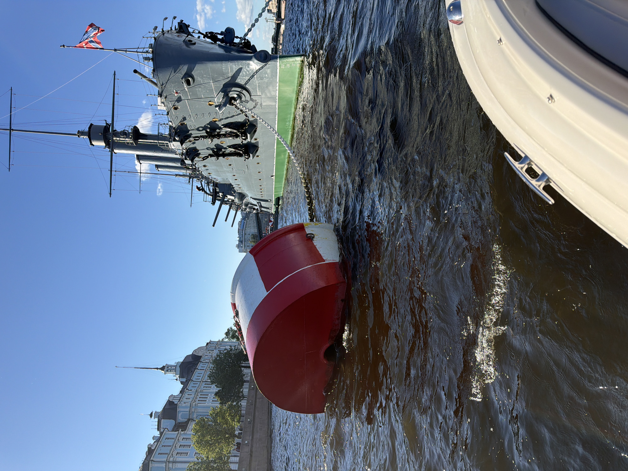 The image shows a warship moored in a river, with a bright red and white buoy floating in the foreground. The warship is painted gray and has several decks and turrets visible. A flag, possibly a naval ensign, flies from a mast. The buoy is cylindrical and has alternating red and white bands. The water is choppy, suggesting a breezy day. In the background, a line of historic buildings with ornate architecture lines the riverbank, suggesting an urban setting. The sky is a clear blue with a few wispy clouds. To the right of the warship, the white hull of a smaller vessel is visible, with a silver cleat attached to its side. The overall scene appears to be a sunny day in a historic port city. The image has been rotated so that the horizon is vertical.