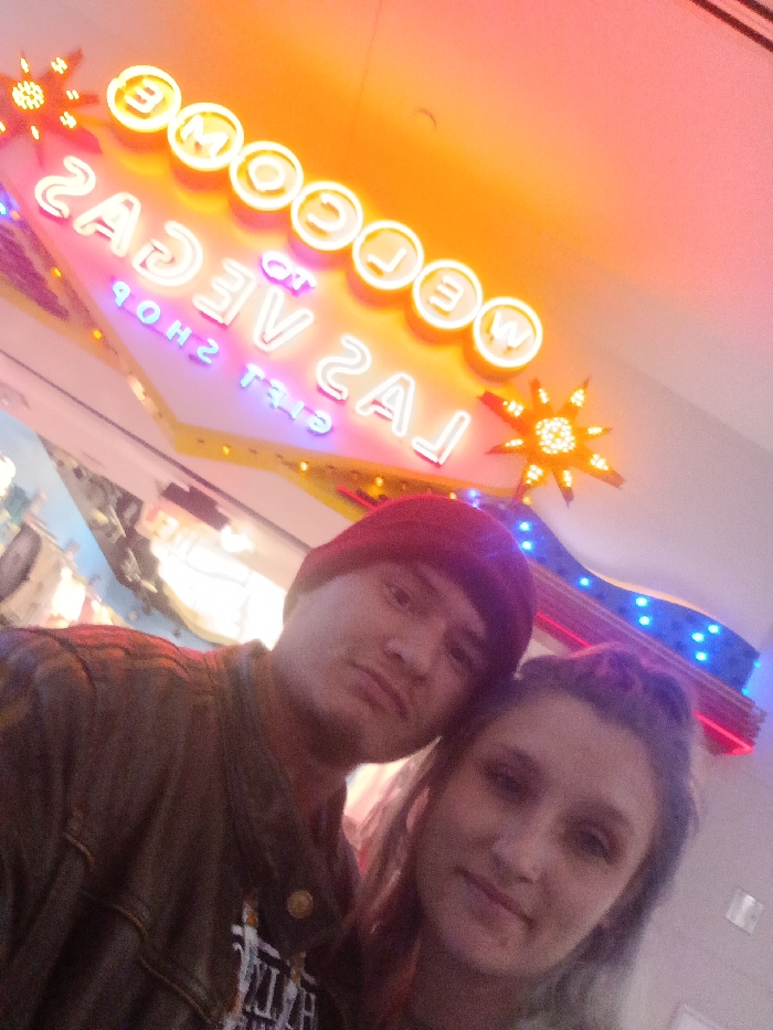 A forward-facing photograph captures two individuals posing together in what appears to be a selfie, illuminated by bright neon lighting. The location is identified as Las Vegas, United States of America, specifically within a gift shop, as indicated by a prominent neon sign in the background displaying the mirrored text "WELCOME TO LAS VEGAS GIFT SHOP" in yellow, pink, and purple neon, flanked by two starburst light elements.
The individual on the left is a male, wearing a red beanie and a dark jacket over a graphic t-shirt. He has a neutral expression, looking directly at the camera. The individual on the right is a female, with light-colored hair, exhibiting a slight smile and also looking directly at the camera. Her head is leaning against the male individual's head. The background shows additional blurred lights and commercial elements consistent with an indoor retail environment.
