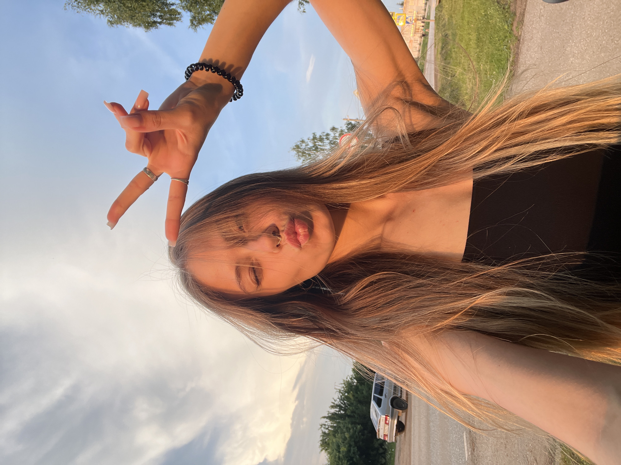 In a candid, selfie-style shot, a young woman with long, blonde hair, possibly in her late teens or early twenties, poses outdoors. She is facing the camera, her eyes are closed, and her lips are pursed as if blowing a kiss. She is wearing a black top. Her right arm is raised, and her hand is held up in a peace sign gesture, adorned with rings and a black beaded bracelet. A faint septum piercing is visible. The background suggests a rural or semi-urban setting. A silver sedan is parked on the side of a dirt road to her left. Lush green trees and a bright, partly cloudy sky dominate the background. The lighting indicates it is either sunrise or sunset, casting a warm, golden hue over the scene. The location is identified as Foky, Russia.