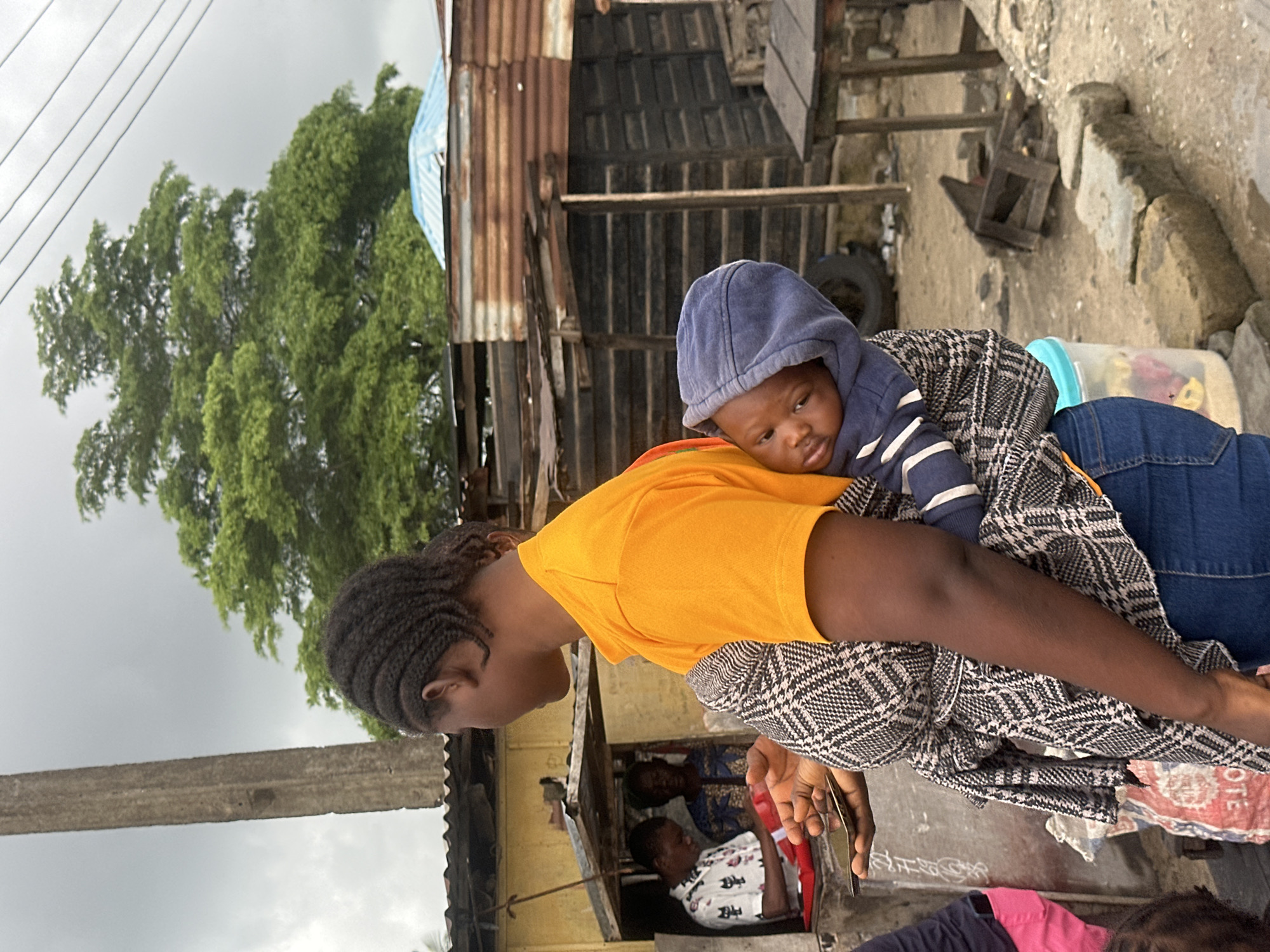 A woman carrying a baby on her back in a patterned wrap stands in what appears to be a market or roadside settlement in Badagry, Nigeria. She wears an orange t-shirt and blue jeans. The baby is wearing a blue hooded sweatshirt and is nestled in the wrap. Behind the woman are several structures made of corrugated metal and wood, suggesting a rustic environment. In the background, a large green tree stands against a cloudy sky. To the left, there is a small stall with people inside. The ground is unpaved and dusty. The overall scene depicts everyday life in a Nigerian community.