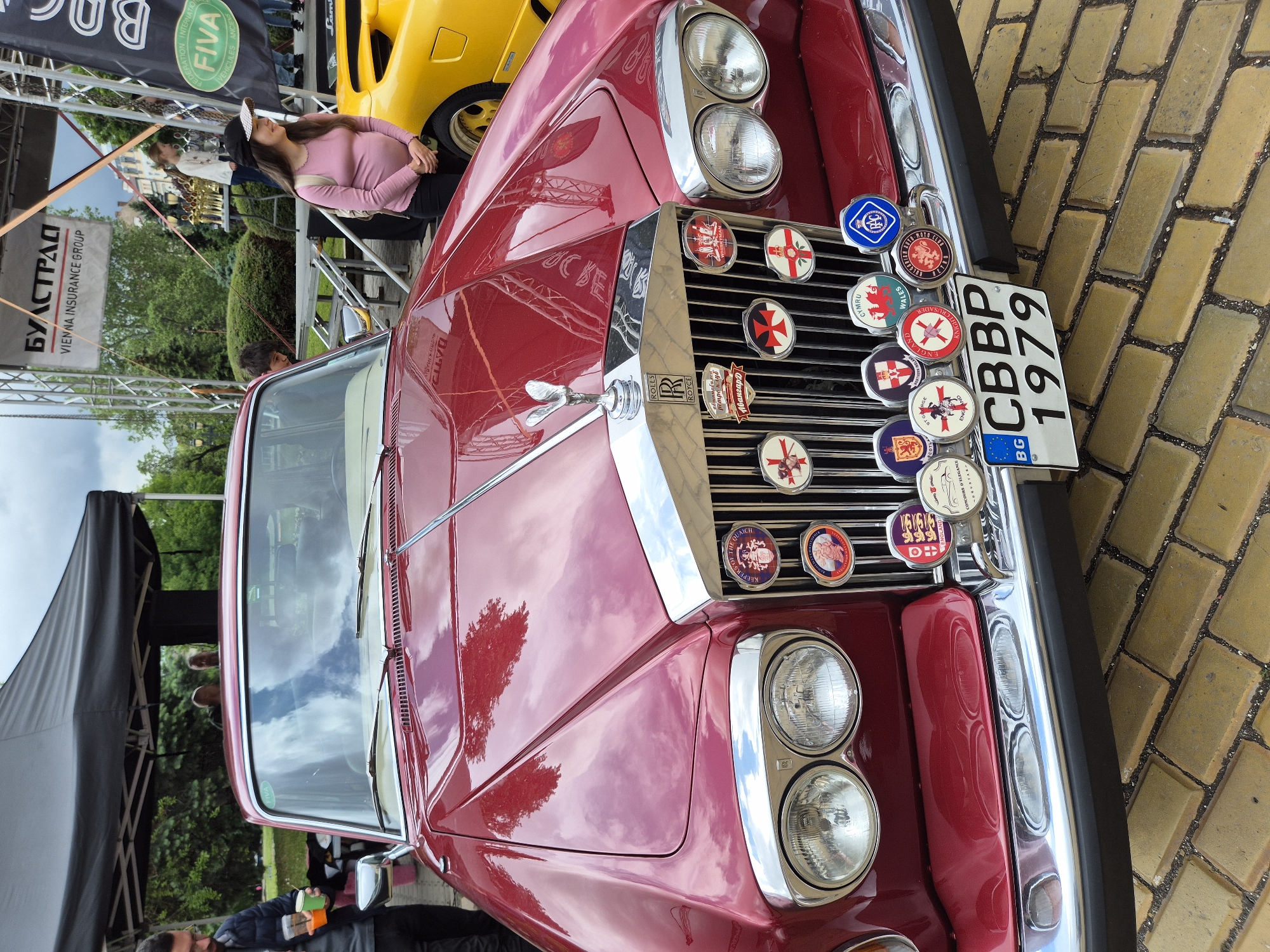 Here is a description of the image:
The image is a ground-level, close-up shot of the front of a vintage red Rolls Royce car. The car is parked on a brick-paved surface. The grille of the car is adorned with numerous circular badges and a Bulgarian license plate reading "CBBP 1979". The hood of the car reflects the sky and some trees.
In the background, a yellow Lamborghini is partially visible. There is also a banner with the words "БУЛСТРАД, VIENNA INSURANCE GROUP" and another banner with "FIVA" and "VEHICULES ANCIENS" on it. A few people are standing in the background near some tables and chairs.
Given the Bulgarian license plate and the "БУЛСТРАД" banner, this image likely was taken in Sofia, Bulgaria.