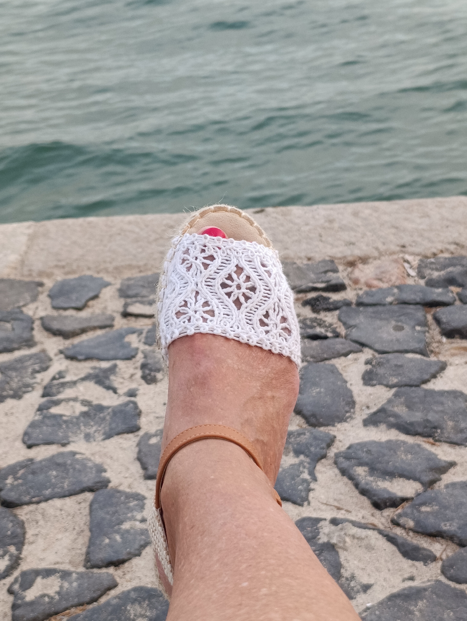 A close-up, first-person perspective shot shows a left foot wearing a white espadrille sandal with a woven lace overlay and a tan ankle strap. The foot is resting on a stone-paved surface, which appears to be part of a promenade or walkway. In the background, a body of water, likely the Sado River estuary, is visible. The stones are varied in size and color, ranging from dark gray to lighter shades, with some sand visible between them. The water is a muted teal color with subtle wave patterns. The overall scene suggests a relaxed outdoor setting, possibly a waterfront area in Setúbal, Portugal, given the characteristic stone paving and proximity to the water.