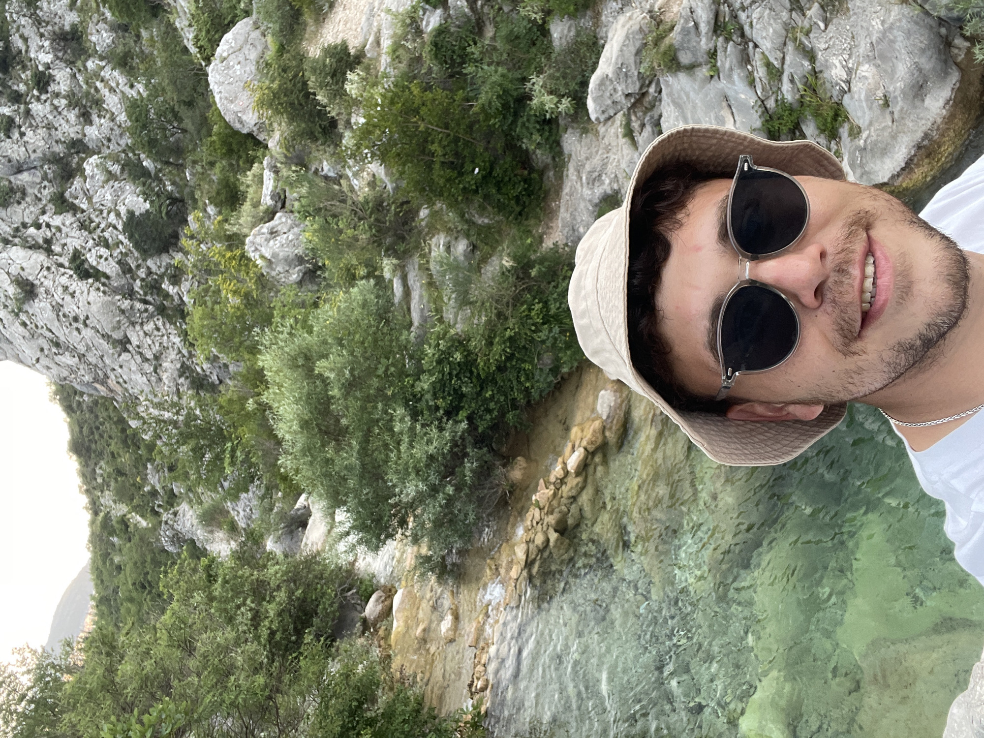 A young man takes a selfie in a scenic natural environment, appearing to be a river or stream flowing through a rocky, vegetated landscape in Zupci, Montenegro. The image is rotated 90 degrees counter-clockwise.
The man, positioned on the right side of the frame, smiles broadly with his mouth slightly open, showing his teeth. He has dark hair and a short beard, and wears dark, round sunglasses that reflect the surroundings. A light beige bucket hat covers his head, and he is dressed in a white t-shirt with a silver chain visible around his neck.
Behind the man, a clear, light turquoise and green body of water, likely a river or stream, winds through the scene. The water reveals a rocky bottom, and its edges are lined with natural stones, some forming a low, rough embankment. The background is dominated by steep, rugged cliffs or mountains, densely covered with varied green trees and bushes that cling to the light grey rock faces. A bright, possibly hazy or overcast sky is visible in the upper left corner, suggesting soft lighting, perhaps during the morning or late afternoon.
The overall scene conveys a sense of natural beauty and tranquility, with the man's smile indicating enjoyment of his surroundings. No visible text is present in the image.