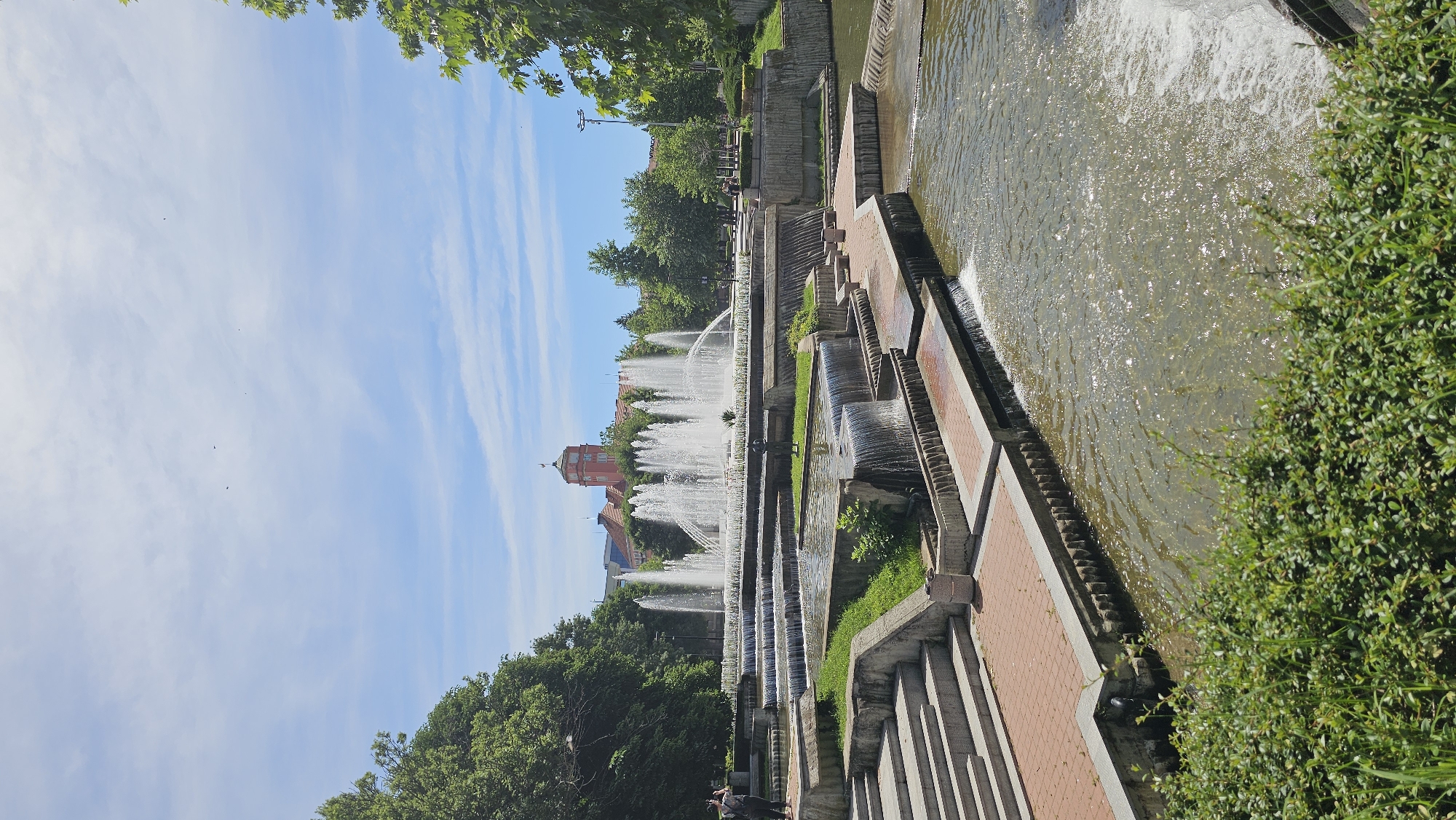 The image captures a water feature in a park-like setting, likely located in an urban area. The water cascades down a series of concrete and brick steps, creating a tiered waterfall effect. Fountains are actively spraying water upwards. Green vegetation, including bushes and trees, is present along the edges of the water feature. A building with a red roof is visible in the background. The sky is blue with streaky clouds.