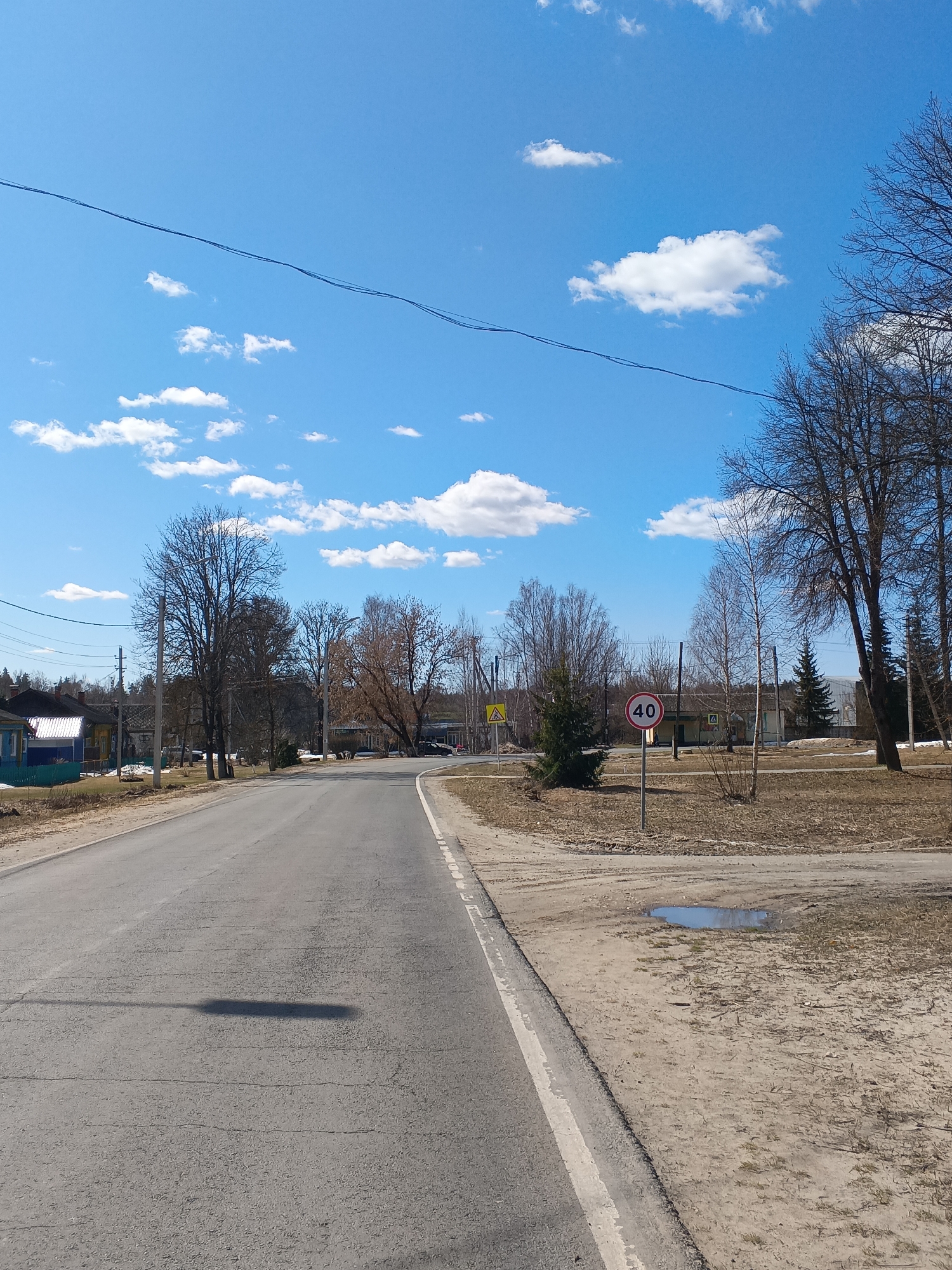 The image depicts a straight section of asphalt road in Красный Маяк, Russia. The road surface has cracks and imperfections. The right side of the road has a sandy shoulder with sparse vegetation and a small puddle of water. A "40" speed limit sign is visible on the right shoulder. Trees line the road, most of which are bare. In the background, buildings and houses dot the landscape. The sky is blue with scattered white clouds and power lines stretching across the top of the frame. The shadow of a lamppost or pole is visible on the road.