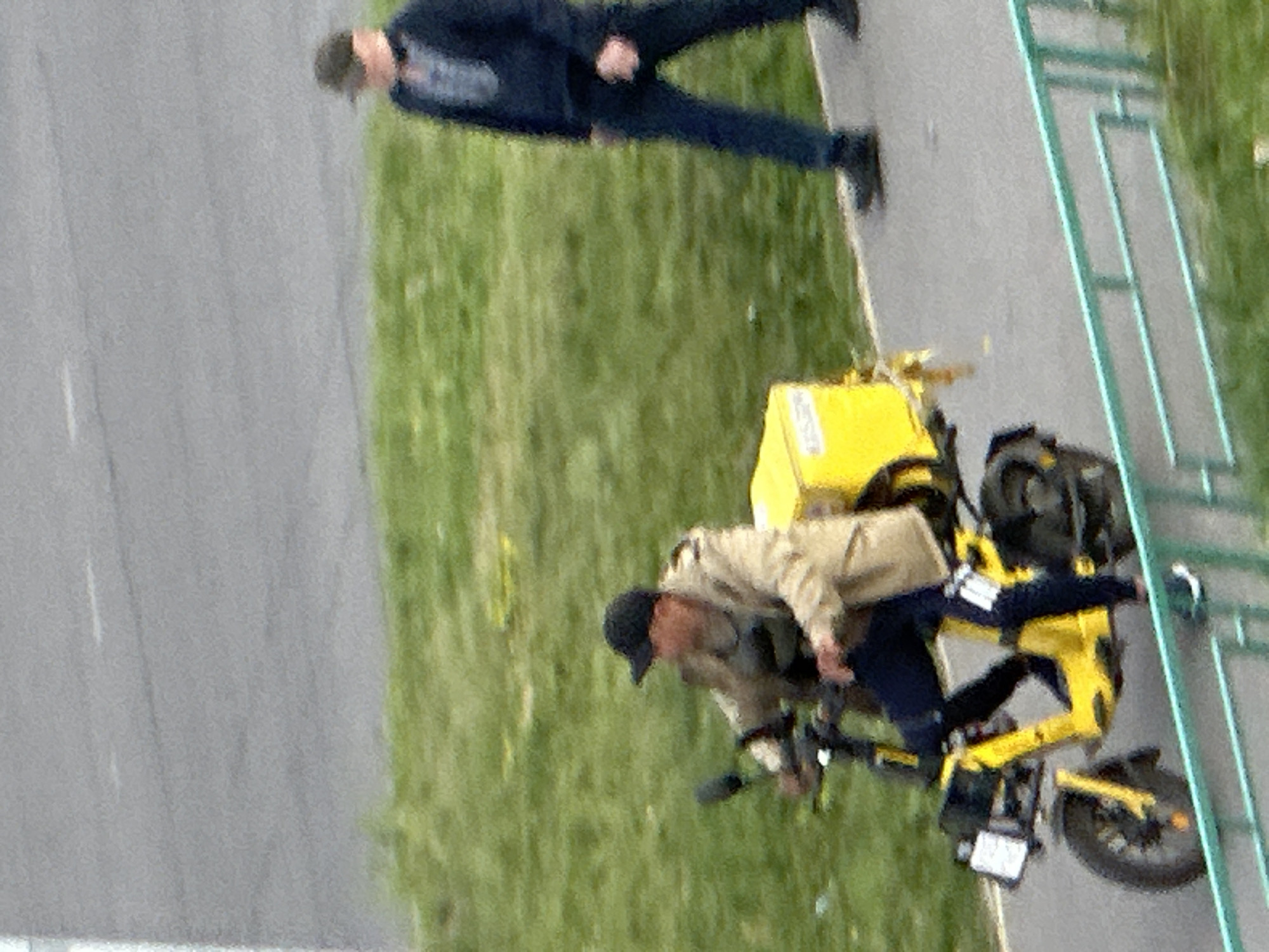 In Podolsk, Russia, a person rides a yellow electric scooter equipped with a large yellow storage box at the rear. The rider, wearing a dark baseball cap, a light-colored jacket, and dark pants, is positioned on the scooter, which has a black handlebar and front wheel. To the right of the scooter, another person walks on a paved surface, dressed in a dark jacket and dark pants, and wearing a dark baseball cap. The background consists of a grassy area adjacent to the paved walkway. A turquoise railing is visible in the foreground, partially obscuring the lower portion of the scooter and the walking individual. The scene appears to be captured outdoors during daylight.