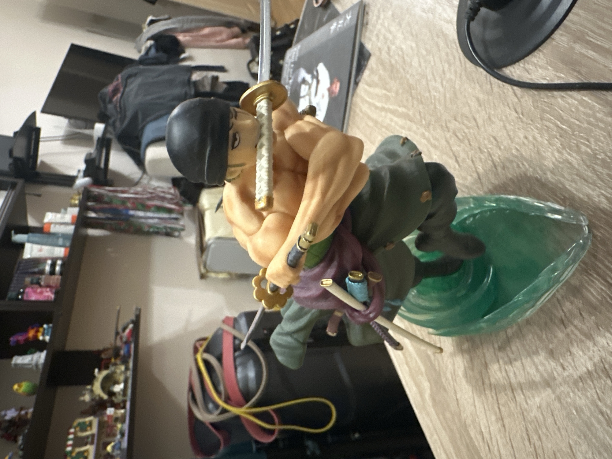 A Roronoa Zoro action figurine is centrally positioned on a light wood-grain table surface. The figurine depicts a muscular male character wearing dark green lower garments, a purple sash, and a black beanie, with two katanas held in his hands and a third clenched in his mouth. He stands on a translucent green base designed to resemble a wave or energy effect. To the right of the figurine, a black circular object with an attached power cord rests on the table.
The background reveals a multi-tiered shelving unit against a light-colored wall. The upper left shelf displays several decorative items, including a yellow-green bird figure, a white Eiffel Tower replica, and various colorful figurines. Below it, the lower left shelf holds multiple miniature structures and objects, some resembling building block sets. On the upper right shelf, a collection of bottles and containers, possibly cosmetics or toiletries, is arranged. Behind the figurine and to the left, a dark cylindrical object is visible, wrapped with yellow and red elastic bands. Further in the background, a flat-screen television is mounted on a wall, with dark clothing draped over unseen furniture below it. The general environment suggests an indoor, domestic setting.
