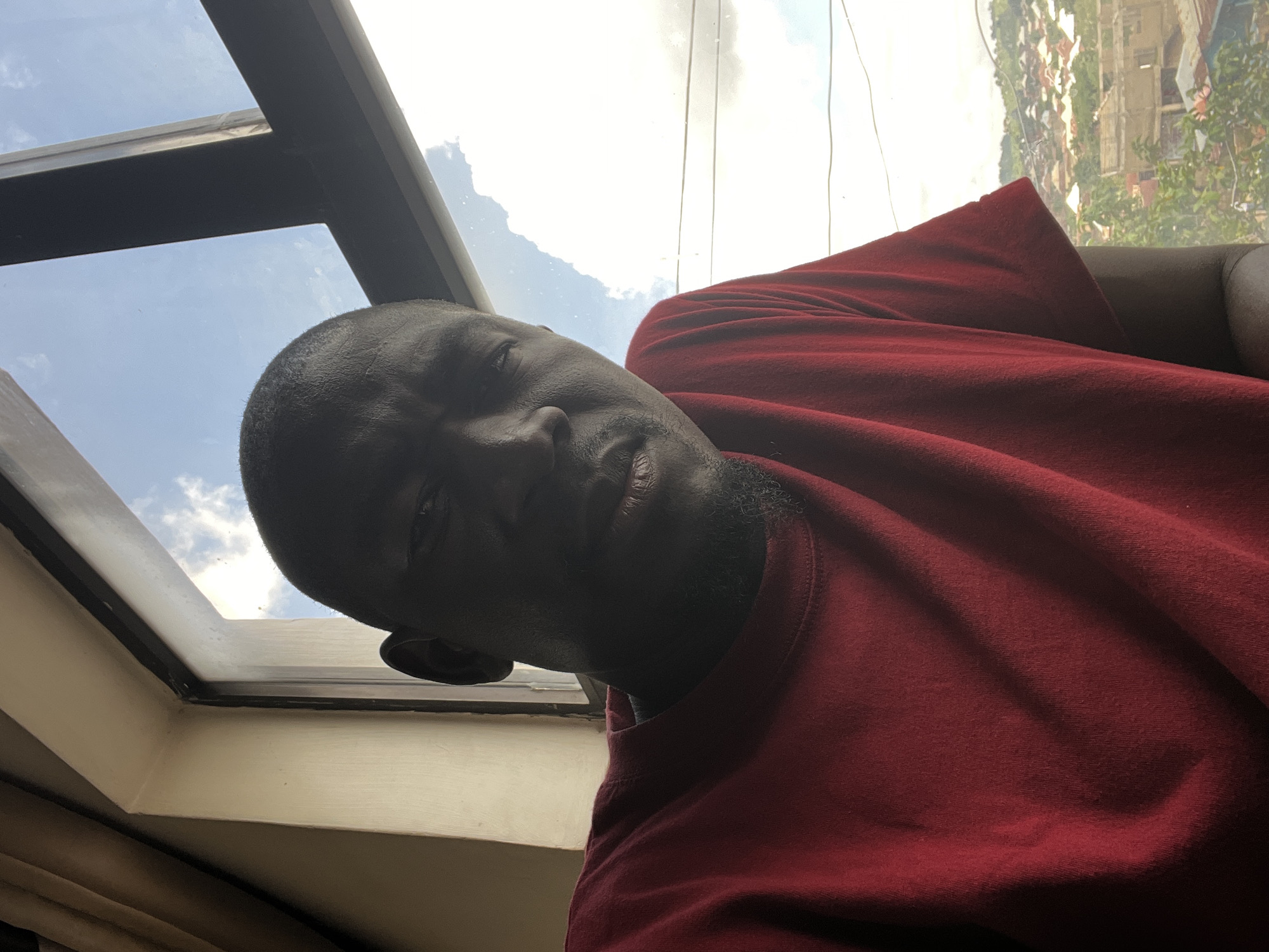 A man is taking a selfie in front of a window. He is wearing a red t-shirt. The window looks out onto a city with houses and trees. The sky is blue with some clouds. The image appears to be taken in the city of Jos, Nigeria.