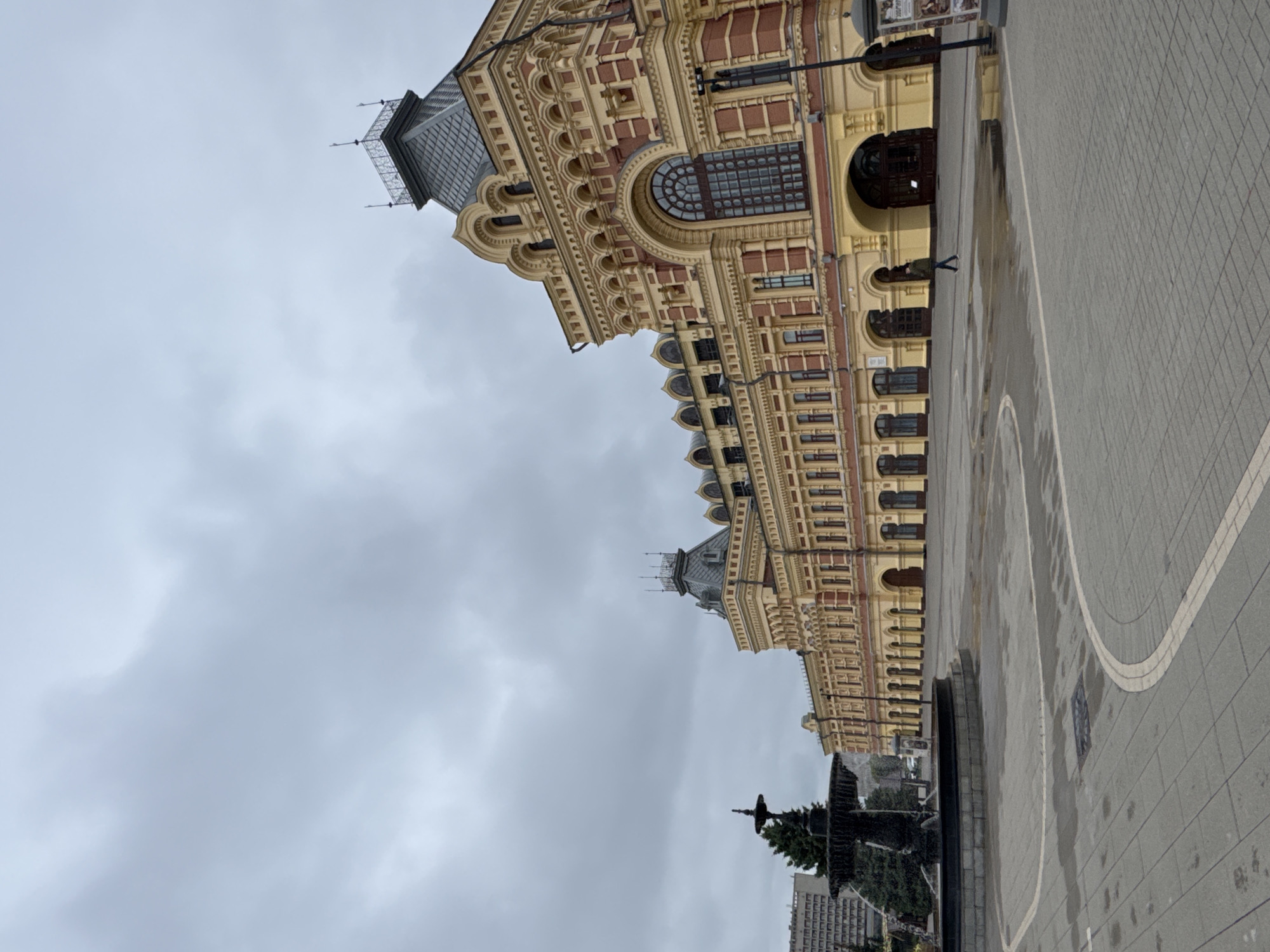 The image shows a low-angle, full shot of the Nizhny Novgorod Fair building, located in Nizhny Novgorod, Russia. The building, painted in vibrant yellow and red, features ornate architectural details, including multiple arched windows and decorative cornices. A prominent dark-colored fountain is situated in the foreground to the left, with water spraying upwards. The ground is paved with gray square tiles, and there are wet patches indicating recent rain. The sky is overcast with a thick layer of gray clouds. A lone individual is walking across the paved area in front of the building, moving from right to left. To the left of the fountain, a tall, dark-colored building with numerous windows is visible.