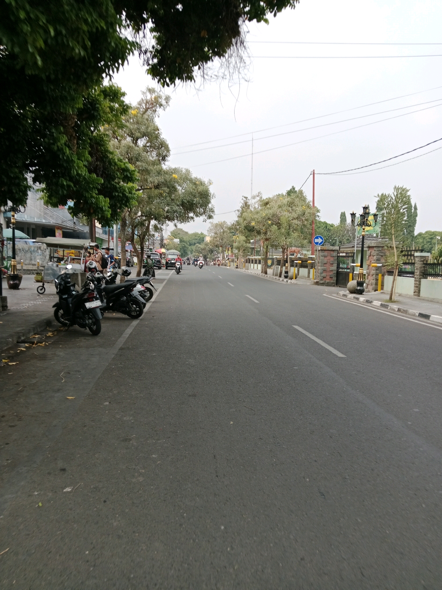 The image depicts a street scene in Cianjur, Indonesia. On the left side of the road, several motorcycles are parked along the sidewalk. A few individuals are standing near the motorcycles. The sidewalk is lined with trees and some structures that appear to be shops or buildings. The road itself is paved and appears to be a main thoroughfare, with traffic visible in the distance, including more motorcycles and cars. On the right side of the road, there is a walled area with decorative lampposts and an entrance gate. The sky is overcast, suggesting a cloudy day. The overall impression is that of a typical urban street in Indonesia during daylight hours.